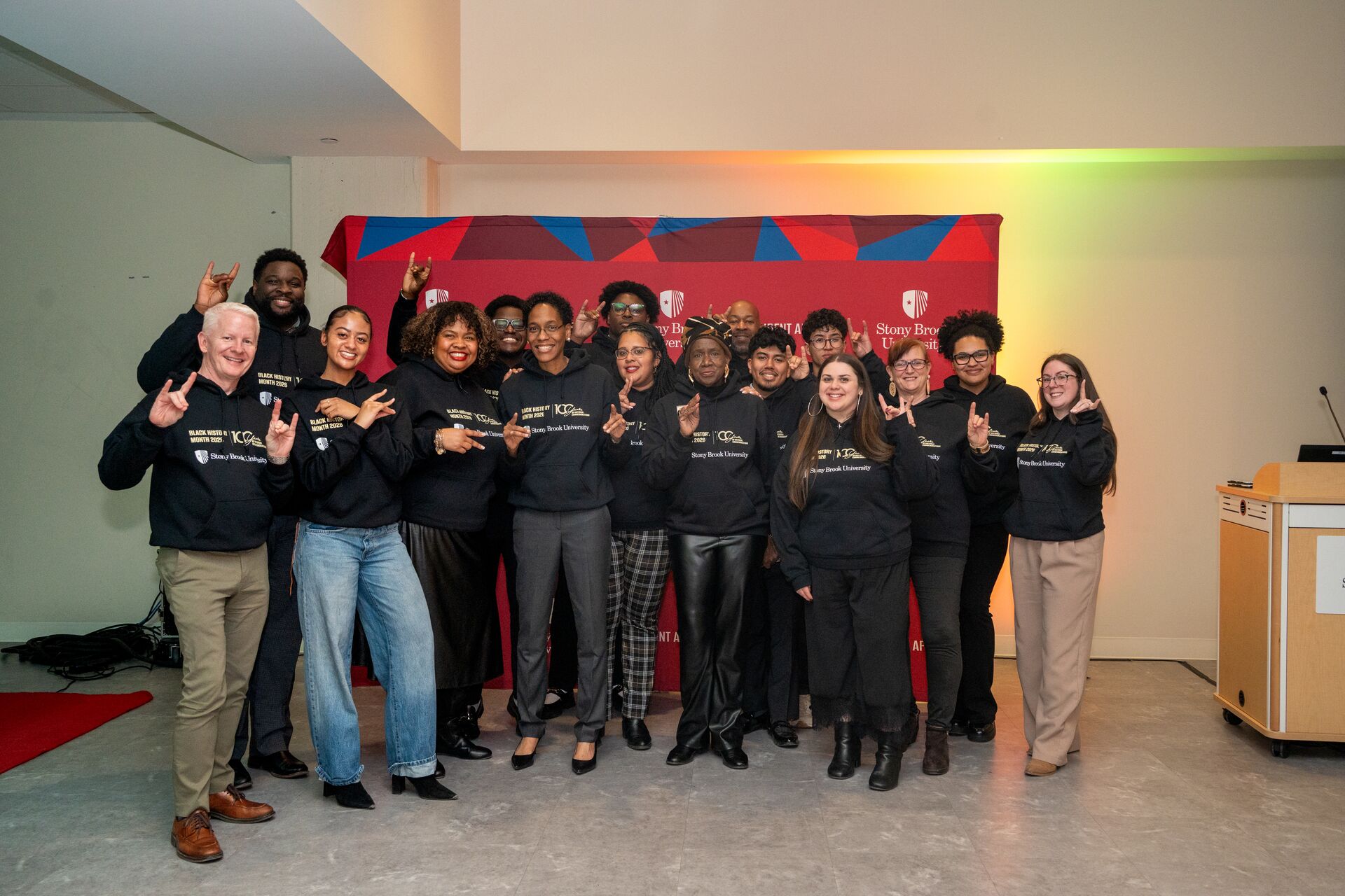 A large group of people stand together indoors for a group photo in front of a red Stony Brook University backdrop. Most are wearing matching black hoodies and smiling while raising their hands to throw up the university's "Wolfie" hand sign.