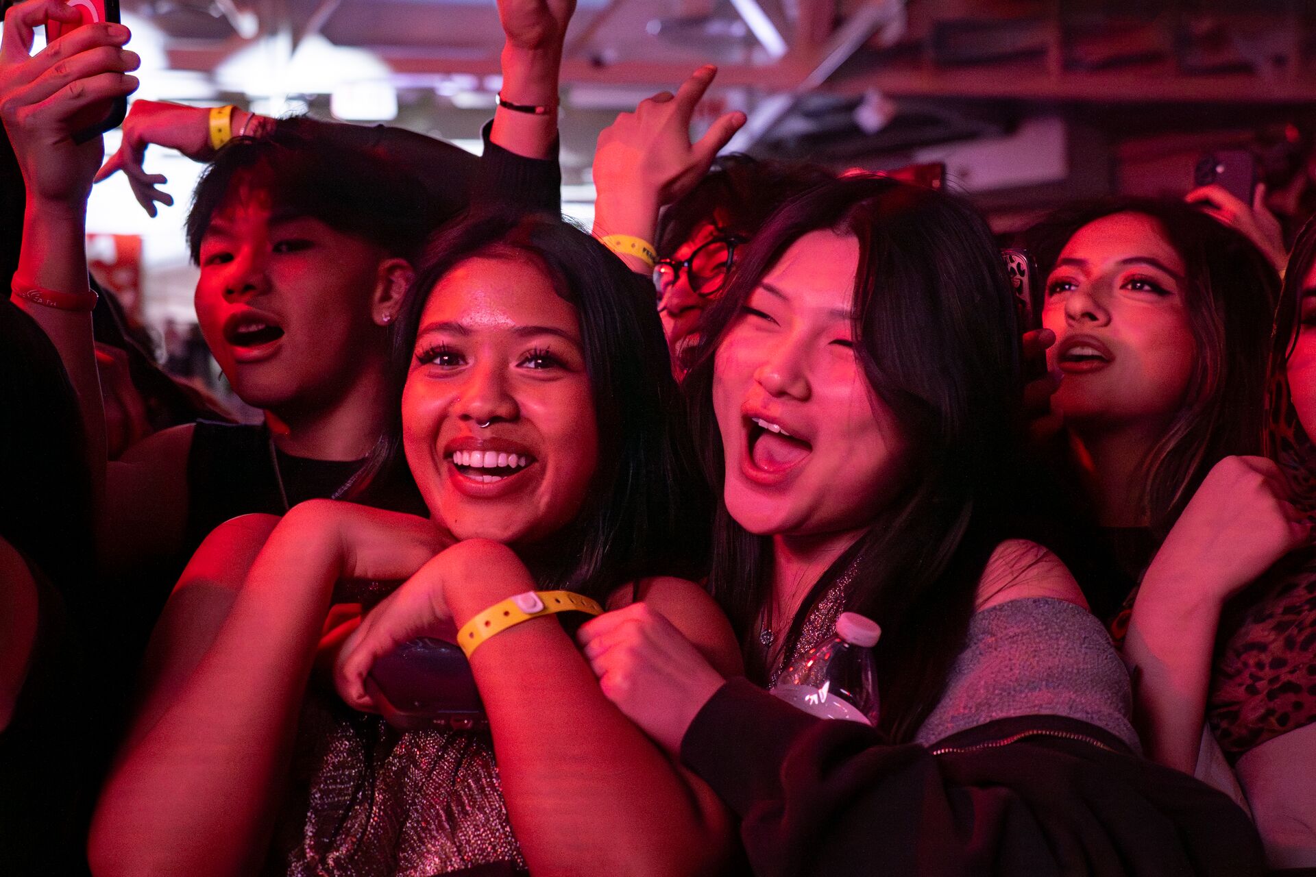 A close-up shot of an excited crowd at a concert, lit by vibrant pink and red stage lights. Two young women in the center front row smile and cheer enthusiastically with their hands raised.