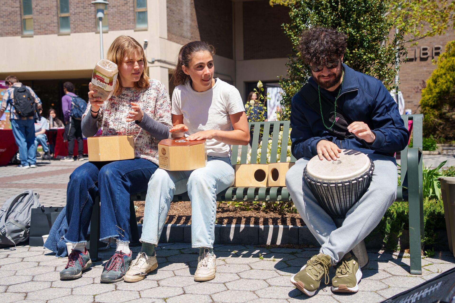 Three young adults sit on an outdoor campus bench playing hand percussion instruments, including two wooden cajons and a djembe drum, during an event with a building and greenery in the background.