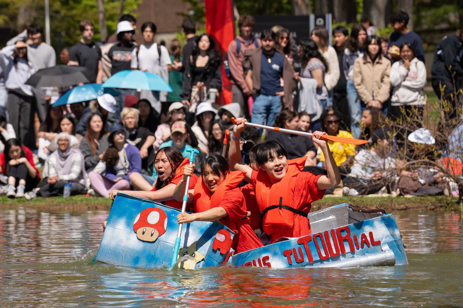 Three students wearing bright orange life jackets eagerly paddle a homemade cardboard boat decorated with Super Mario motifs across a pond, while a large crowd of spectators watches from the grassy bank behind them.
