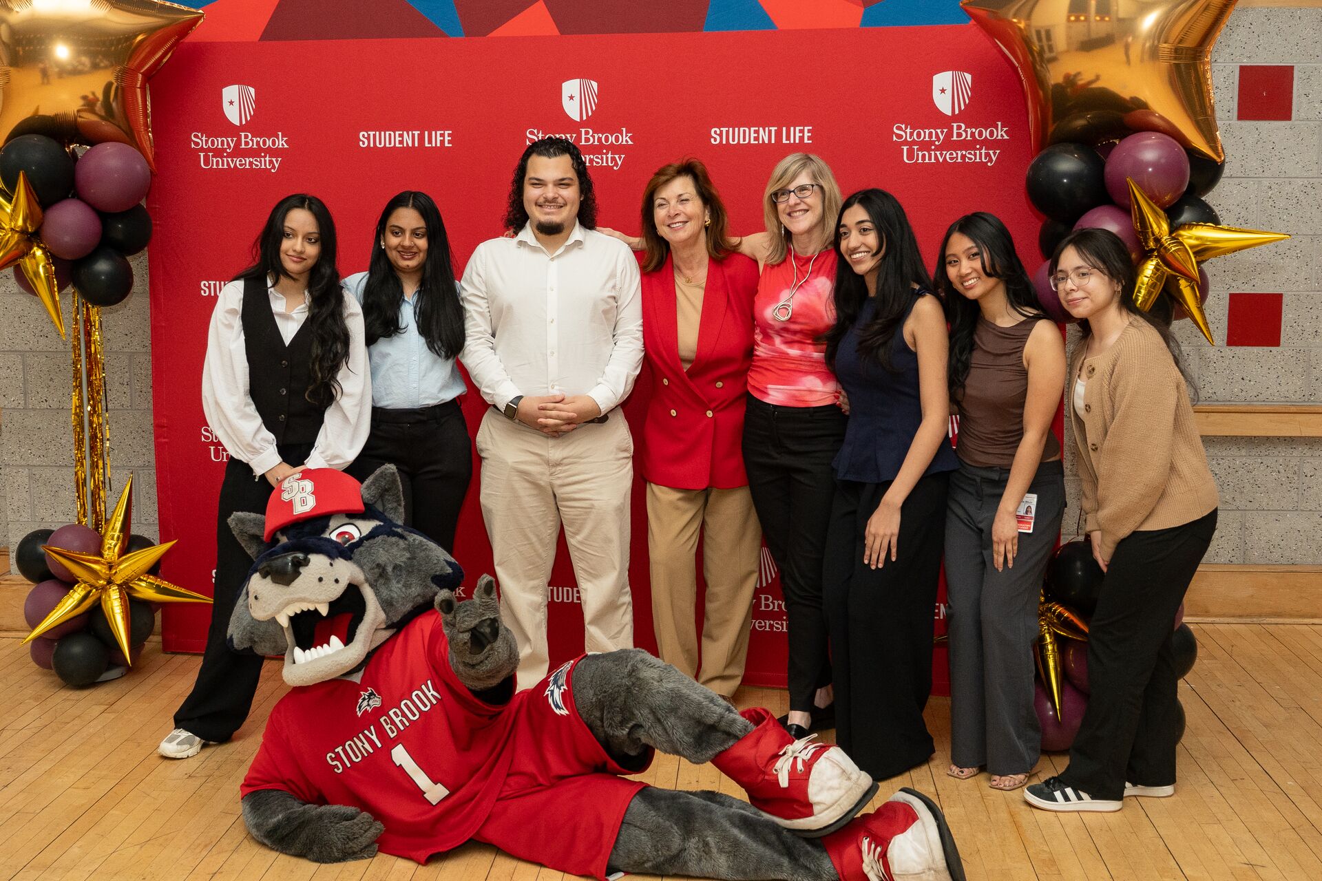 A group of seven students and administrators smile together in front of a red Stony Brook University backdrop. In the foreground, the university's wolf mascot lies on the wooden floor, propped up on an elbow and waving.