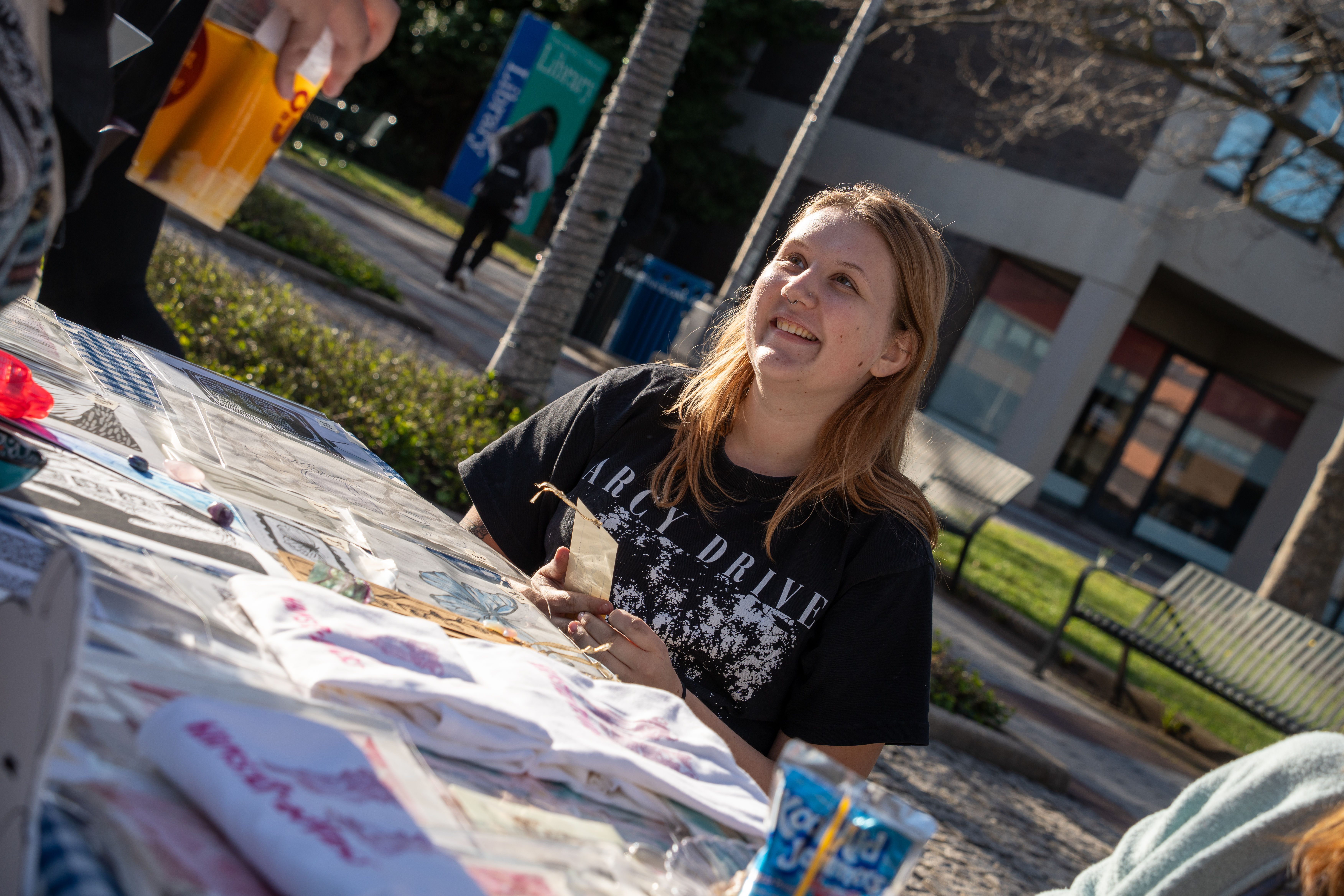A smiling student in a black graphic t-shirt sitting behind a display table with merchandise during an outdoor campus festival.