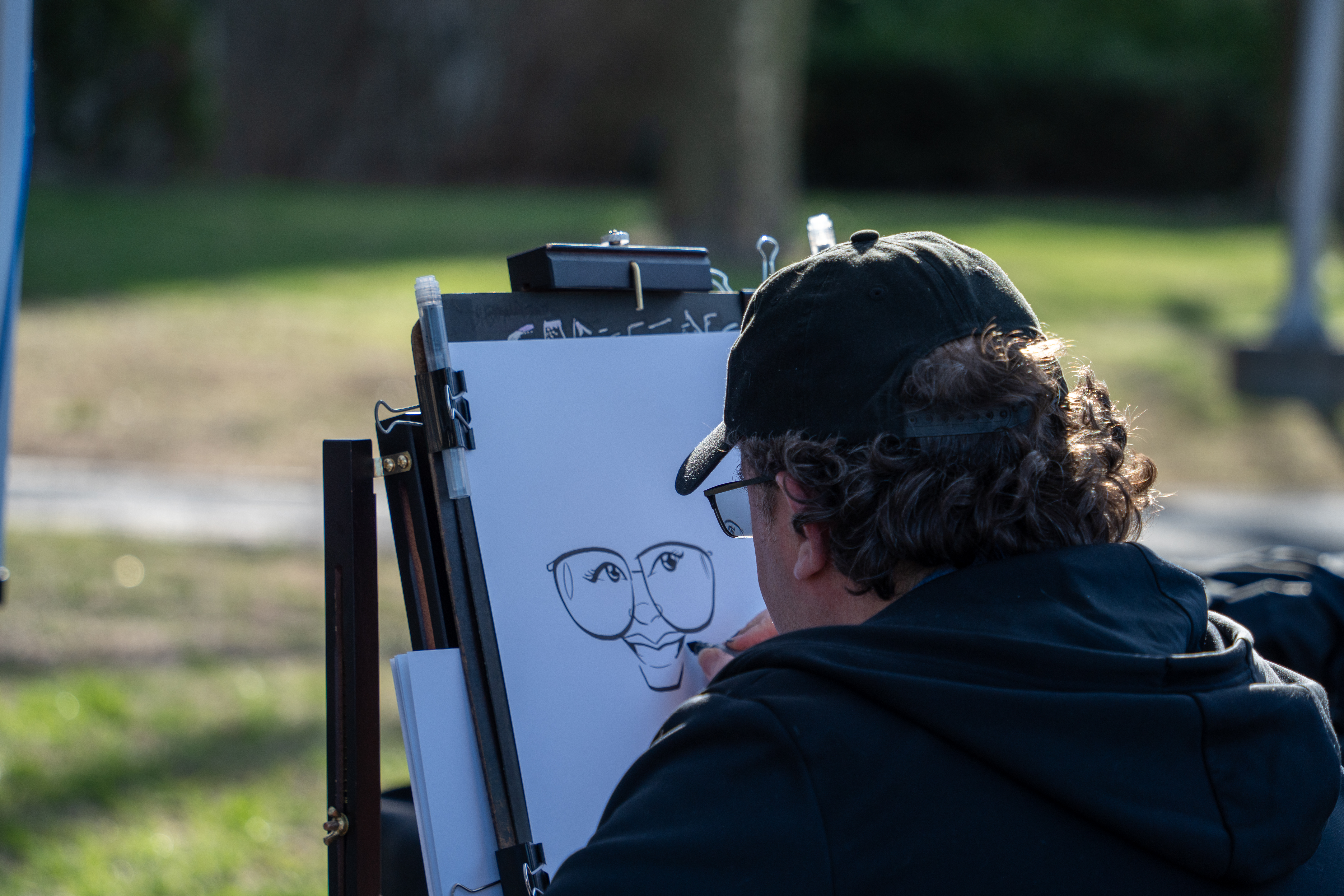 An artist from behind wearing a black baseball cap, drawing a black-and-white caricature of a face with large glasses on an easel outdoors.