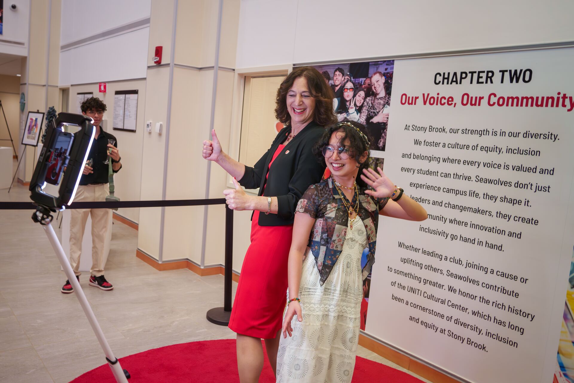Two women smiling, giving thumbs up, and waving in front of a 360-degree ring light photo booth setup next to a Stony Brook University informational sign.