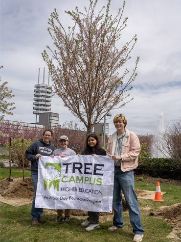 Staff and students holding Tree Campus USA banner in front of newly planted tree