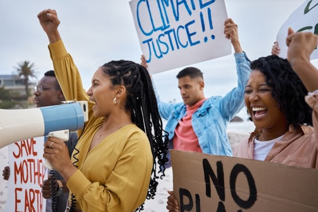 A group of diverse protesters stand outdoors near a beach, raising their fists and holding signs. One person speaks into a megaphone while others smile and chant. Visible signs read “Climate Justice!!,” “No Planet B,” and “Save Mother Earth,” conveying a passionate climate activism demonstration.