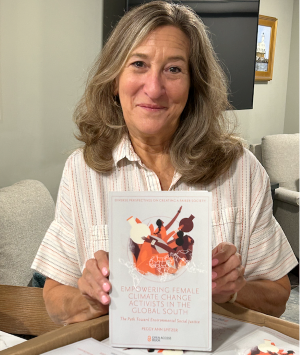 Peg Spitzer smiling indoors holds up her book titled Empowering Female Climate Change Activists in the Global South: The Path Toward Environmental Social Justice.