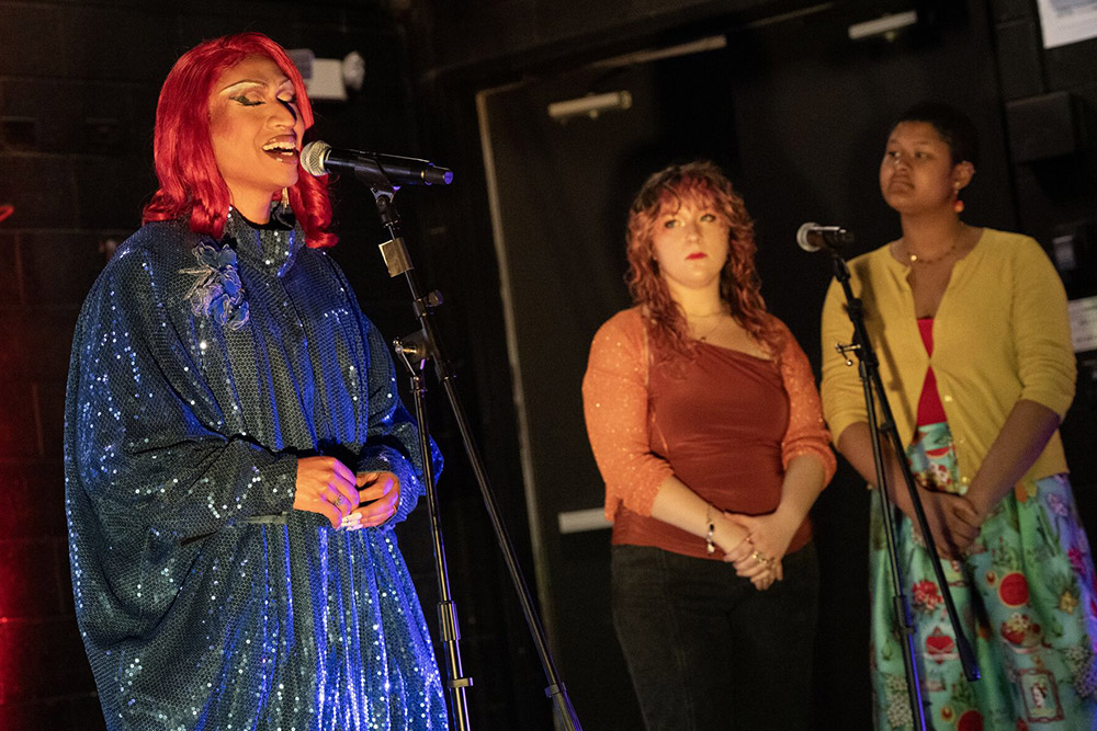 Gad Yola performs at a microphone in a blue sequined gown alongside two students during her residency at Stony Brook University.