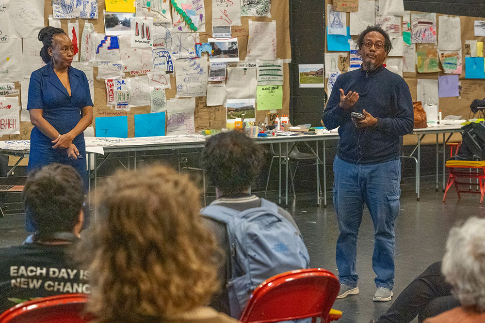 Two presenters address an audience in a studio space with student artwork covering the walls.