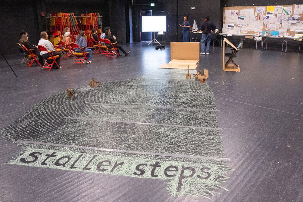 Audience seated on colorful chairs during a presentation in a black box theater, with "Staller Steps" written in chalk on the floor.