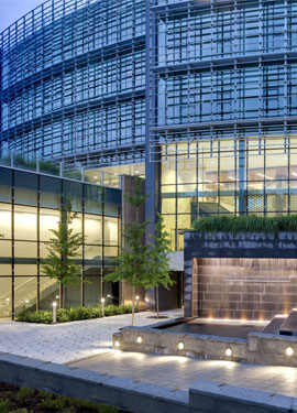 Glass-fronted university building with illuminated interior spaces, landscaped walkway, and water feature in the foreground.