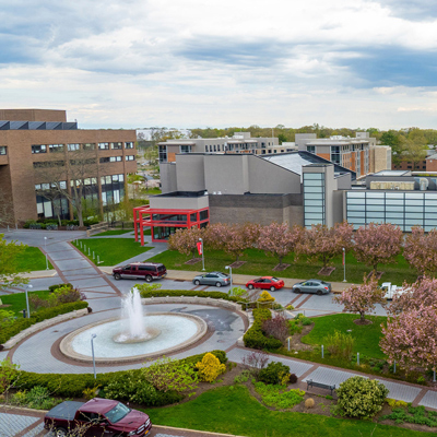 An aerial view of Stony Brook University campus featuring a circular fountain in the foreground. Pink flowering trees line a paved walkway where several cars are parked. Modern brick and glass academic buildings stand under a cloudy sky.