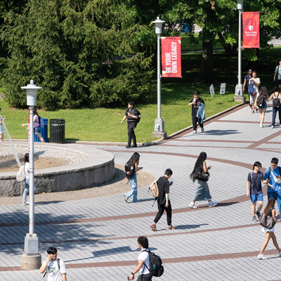 A high-angle shot of a sunny Stony Brook University plaza where students are walking across a paved, patterned walkway. Red banners with white text, including "BE YOUR OWN LEGACY," hang from lamp posts. In the background, lush green trees and a stone fountain edge are visible.