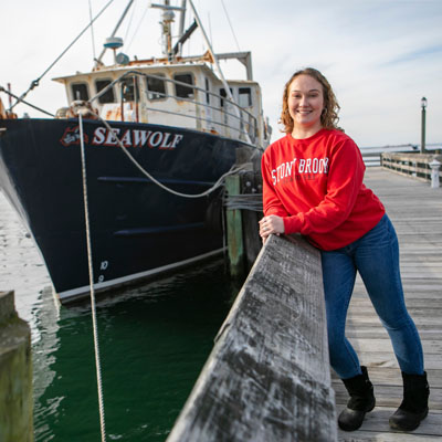 A woman leans against a pier with the Seawolf, Stony Brook's research vessel, behind her.