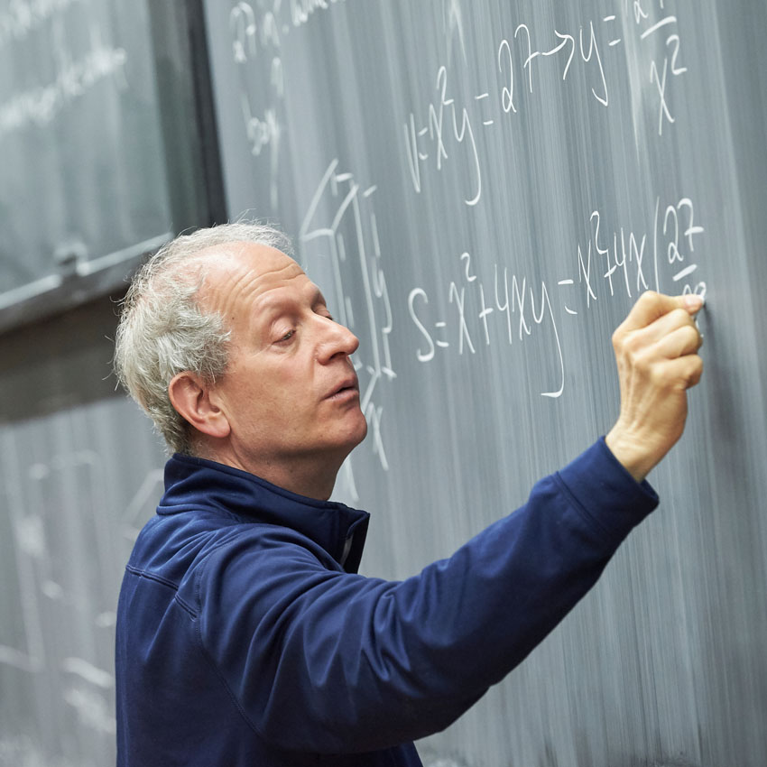 Male professor wearing a navy shirt writes an equation on a blackboard