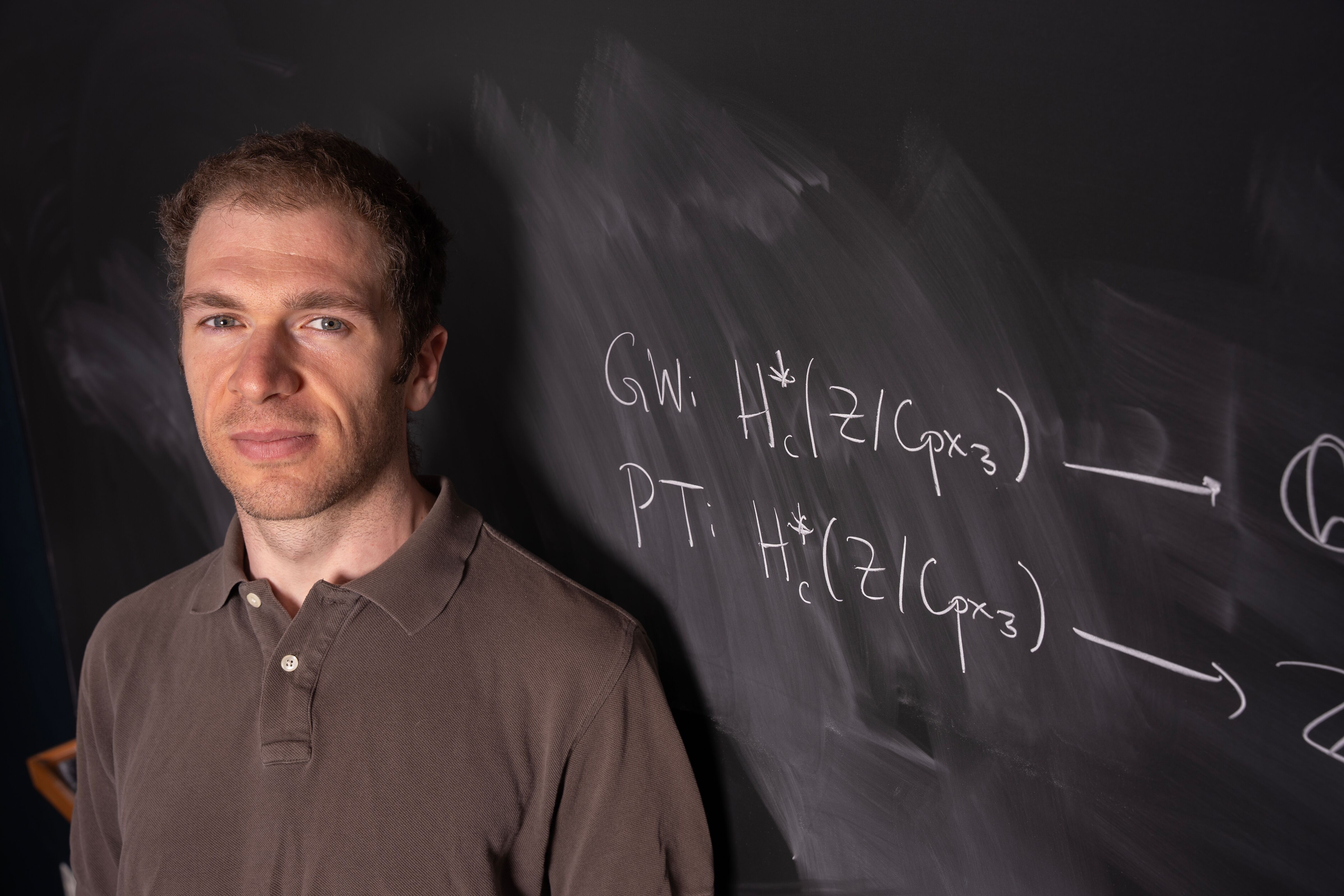 Male professor wearing a brown shirt stands in front of an equation on a blackboard