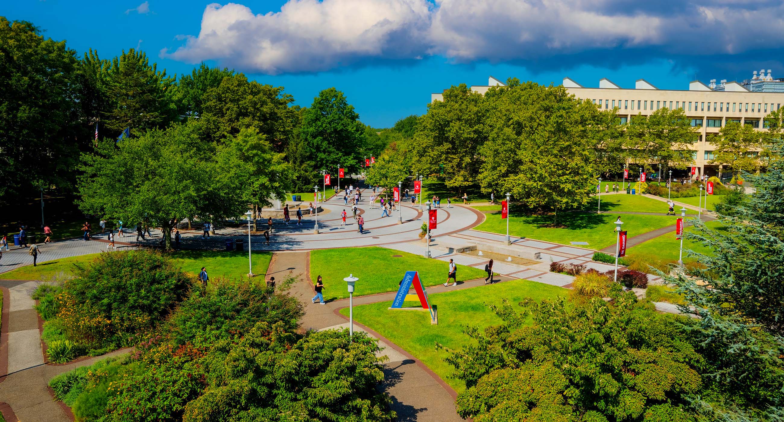 A busy walkway on a tree-lined university campus with students and motivational banners.
