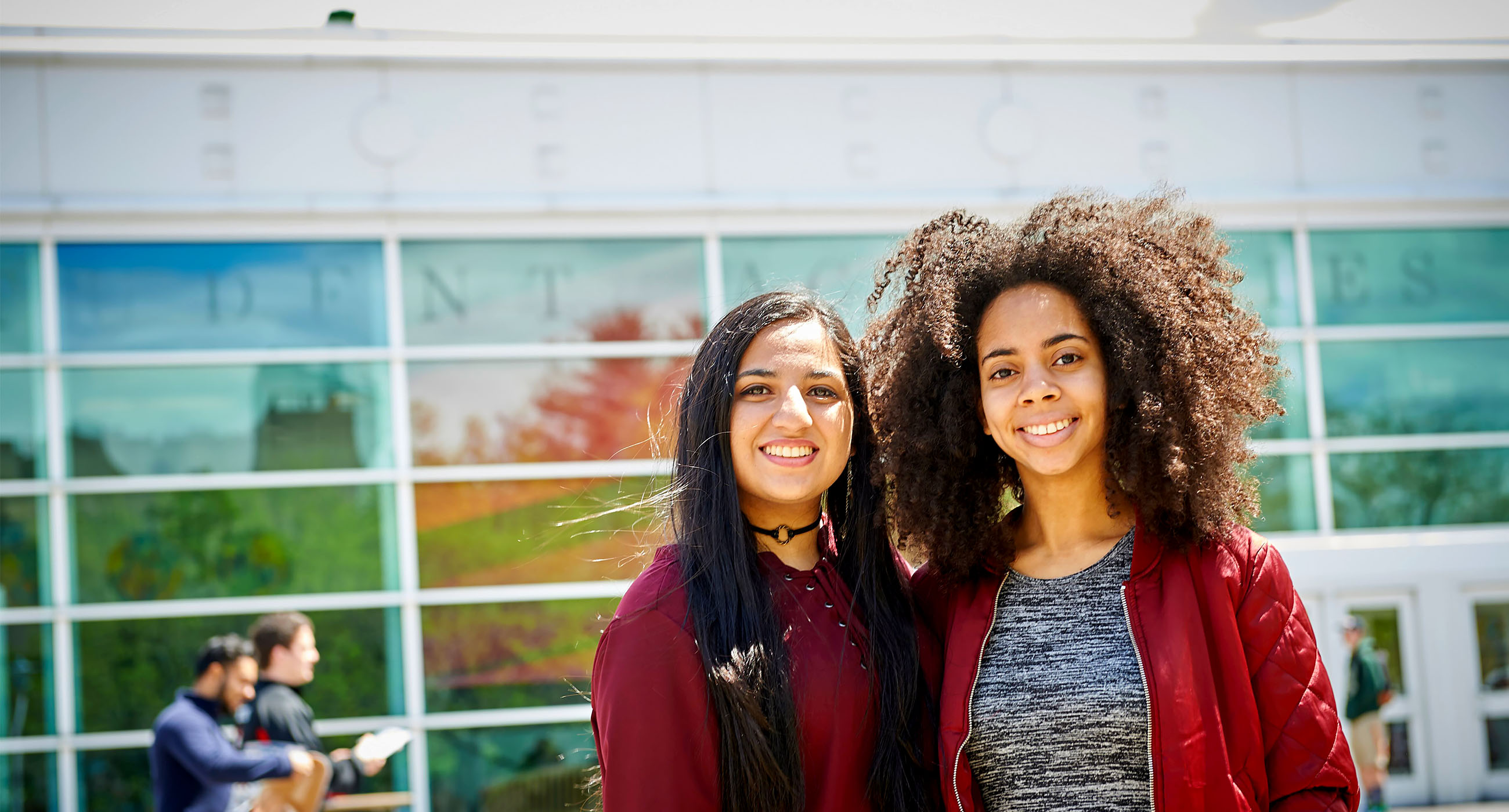 Two students posing for a picture smiling outside of the Student Activities Center at Stony Brook University