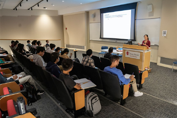 Students sitting in auditorium style chairs in a classroom with professor standing at a podium presenting