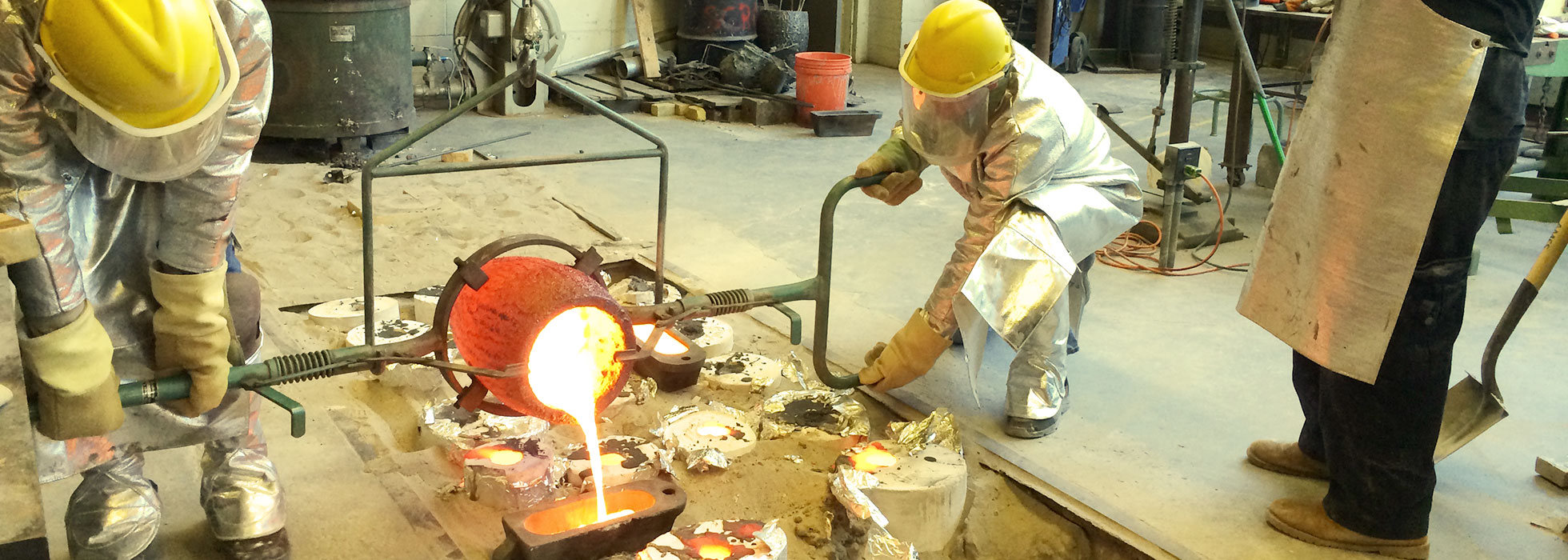 An instructor and a student, clad in heat-resistant suits, engaged in bronze casting work.