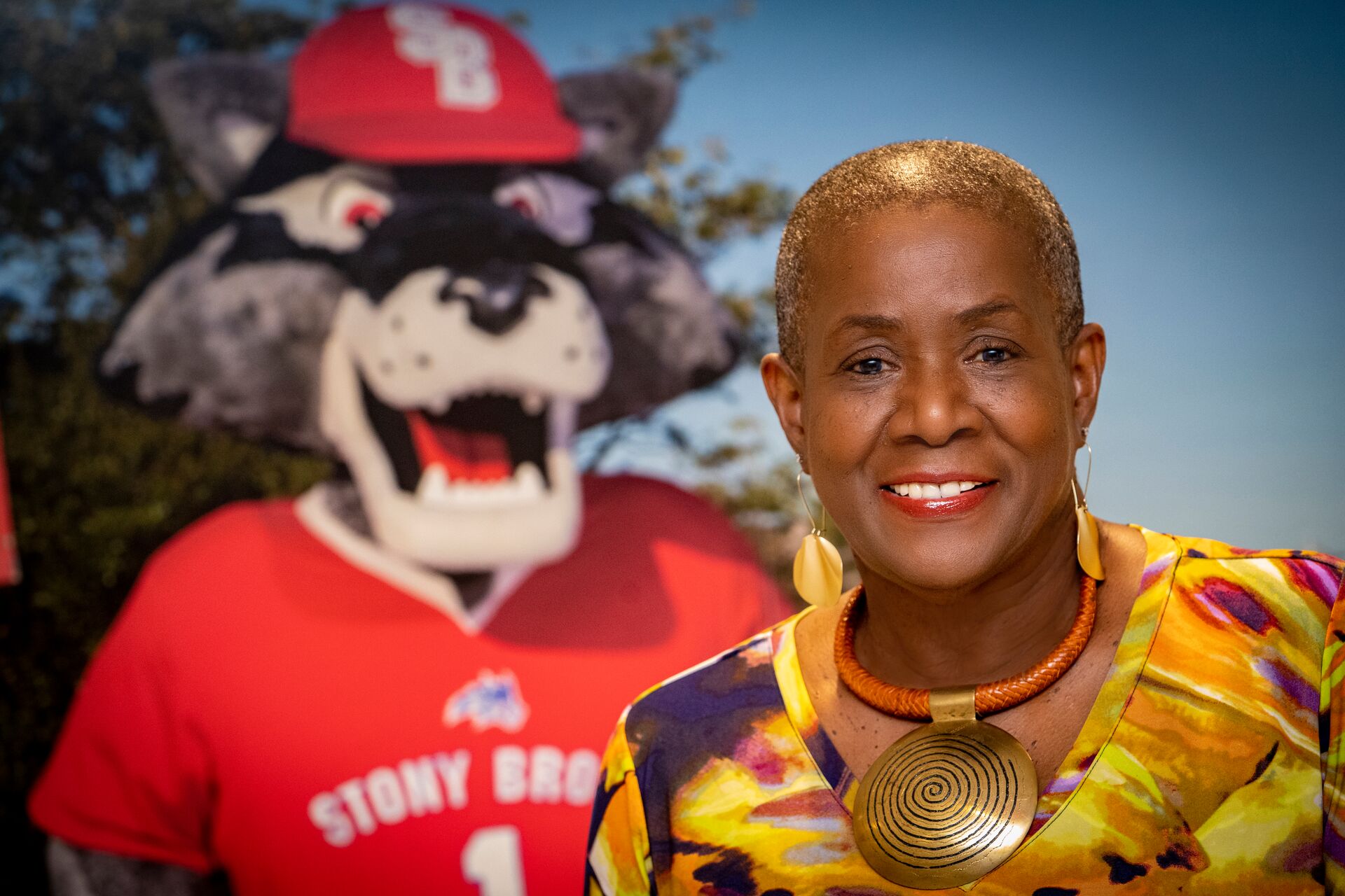 Older African American woman with closely shaved head and blonde hair growing back, wearing a yellow and orange multicolored swirls shirt, large leaf shaped yellow earrings, and a large gold medallion necklace; standing in front of a cutout of the Stony Brook University mascot Wolfie