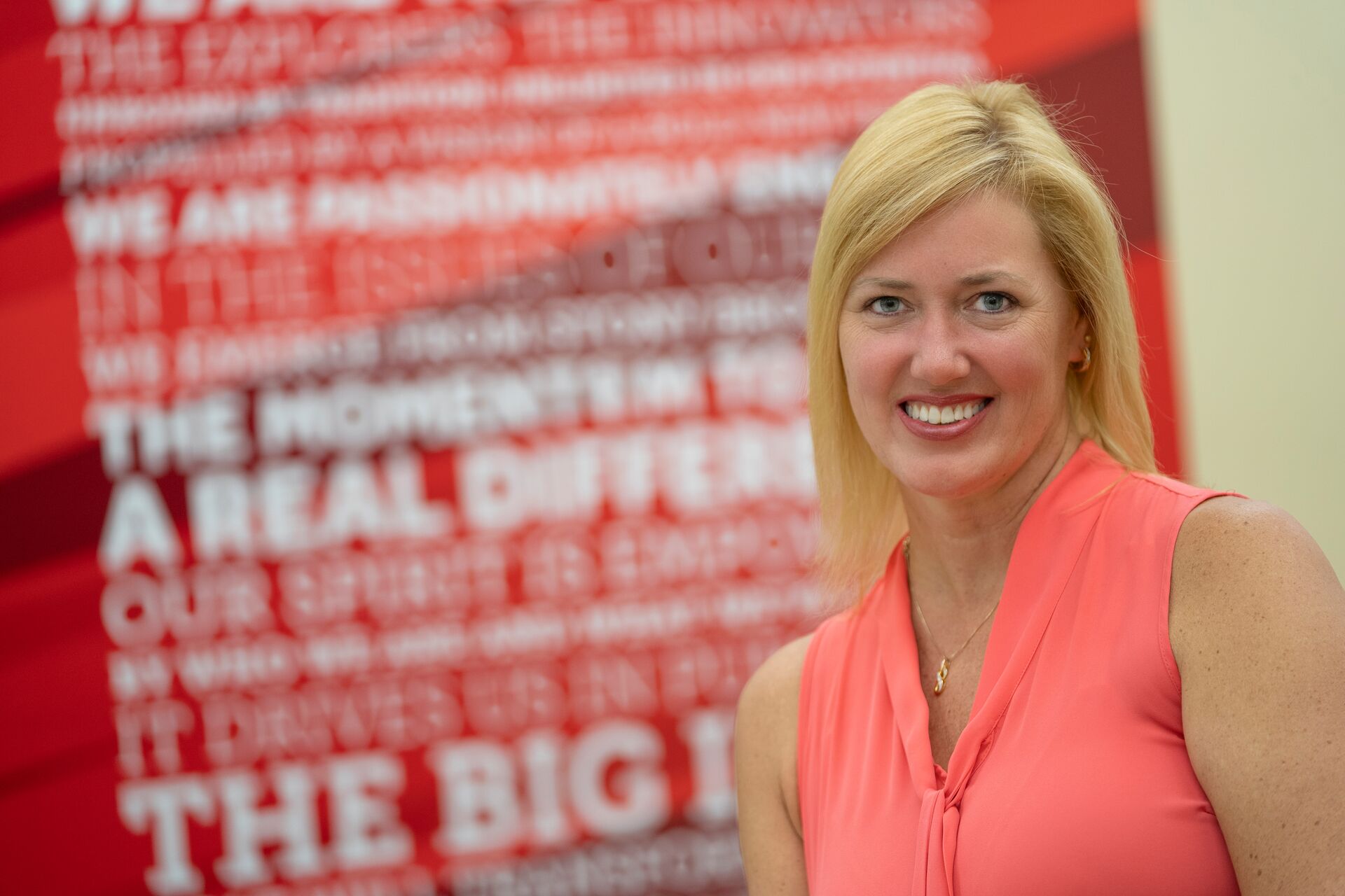 Blonde caucasian woman wearing sleeveless salmon blouse standing in front of wall with red and white blurred words