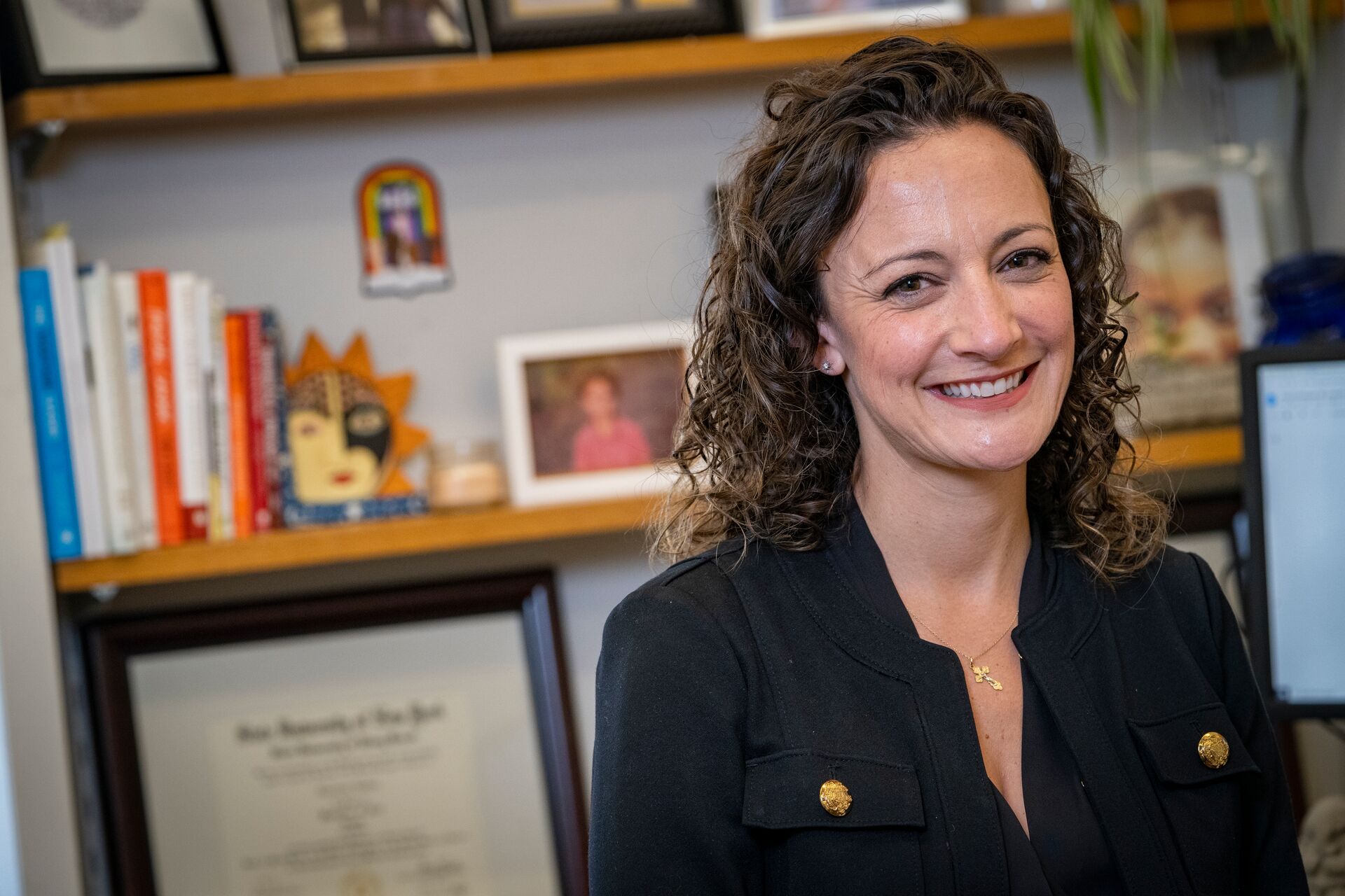 Caucausian woman with curly brown mid-length hair, wearing black shirt with gold buttons on pockets, gold cross necklace, standing in front of a wall with two shelves filled with books