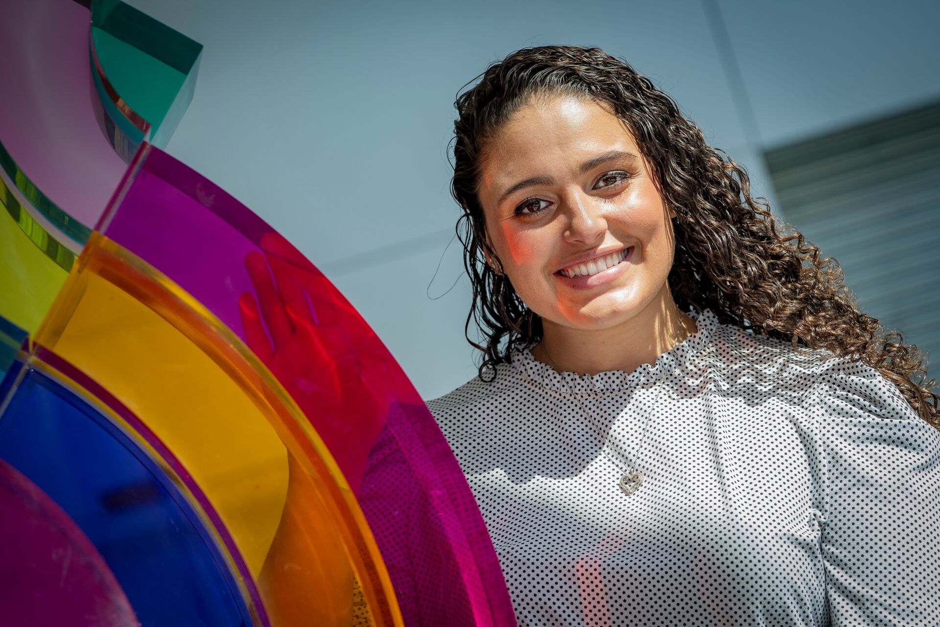 Woman with long curly hair wearing shirt with high neck and standing next to rainbow acrylic statue