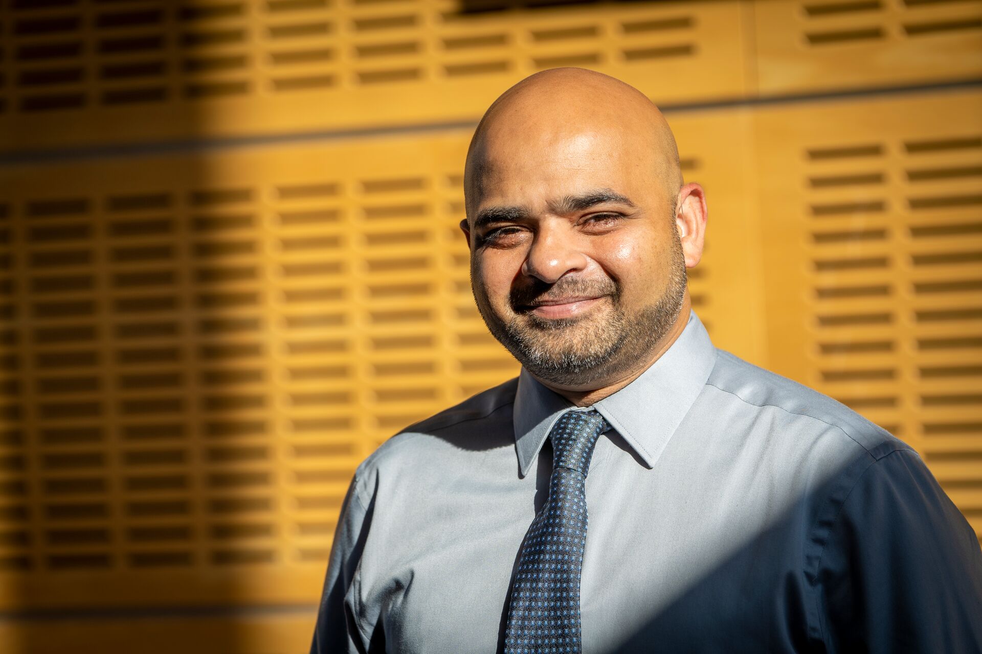 Southeast Asian/caucasian man with scruff, blue button down and tie, leaning against textured wall