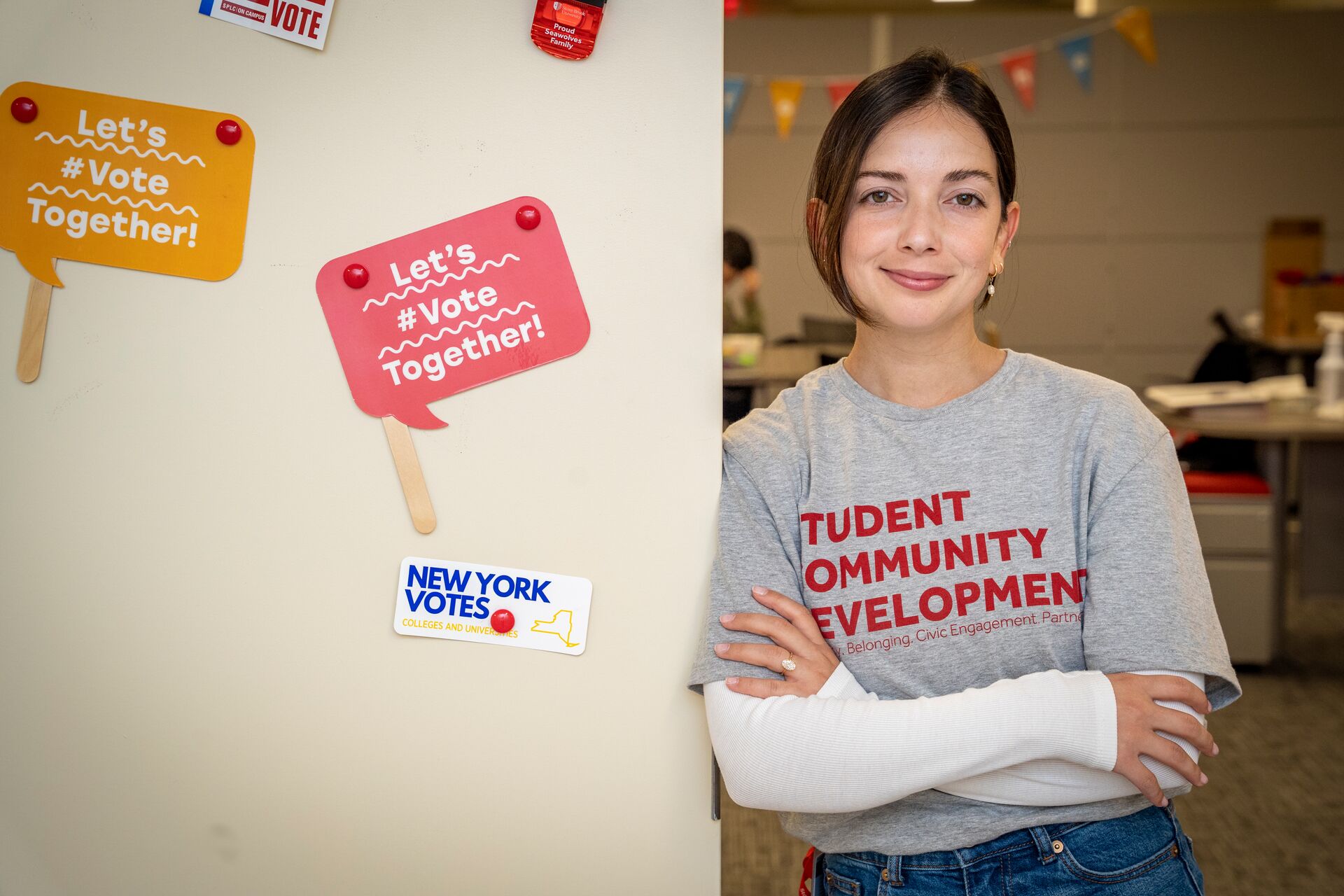 Caucasian hispanic woman with brown hair pulled back, wearing a gray t-shirt over a white t-shirt - the gray t-shirt says, "Student Community Development;" the woman's arms are crossed. She is leaning against a wall with two signs (one orange, one pink) that say, "Let's #vote together." There is also a sticker on the wall that says, "New York Votes"