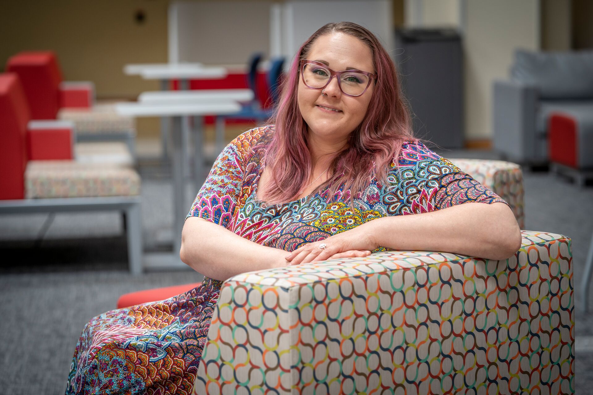 Caucasian woman with long pink hair and glasses wearing multicolored short-sleeve dress, sitting in a gray chair with multicolored circles on it