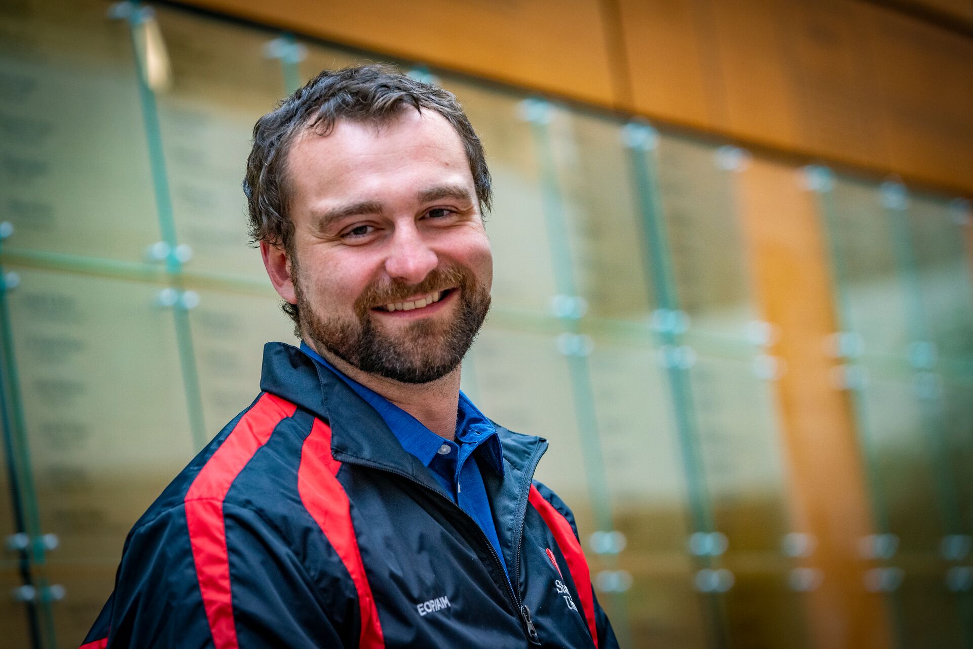 Caucasian man with dark short hair and facial ahir, wearing blue and red athletic shirt