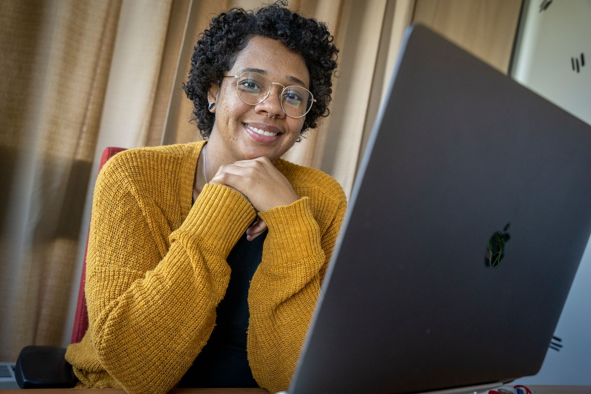 African American woman with shorty curly hair and glasses, hands clasped below chin, wearing golden rod cardigan over black shirt, desktop monitor facing her