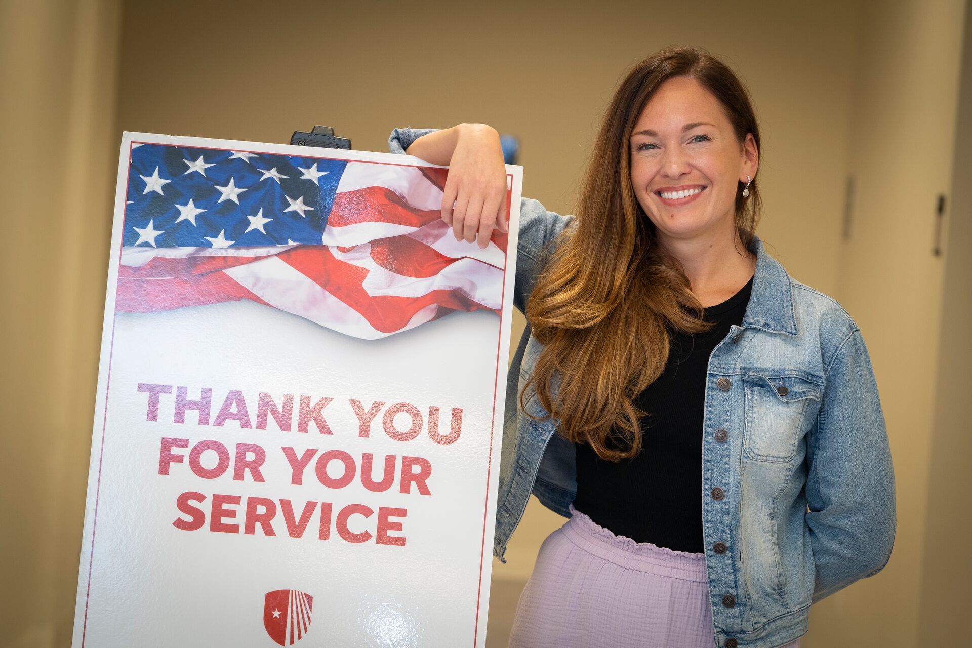 Caucasian woman with long chestnut hair wearing jean jacket over black shirt tucked into lavendar skirt, standing with arm over a sign that says, "Thank you for your service."