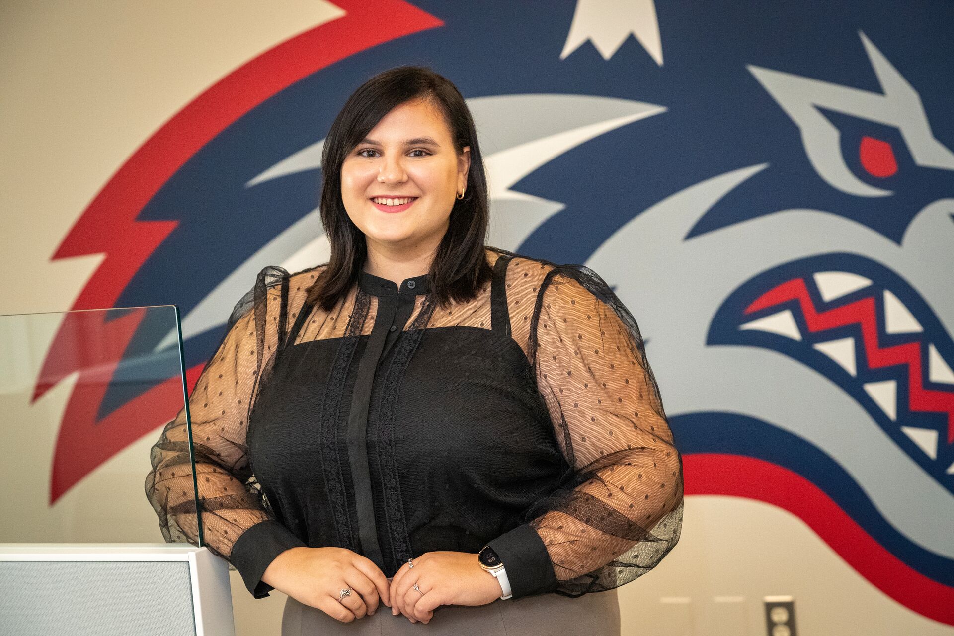 A woman (Rachael Eyler), with dark hair, wearing a black sheer-sleeved blouse, standing in front of a Stony Brook University wolfie logo mural in the background.