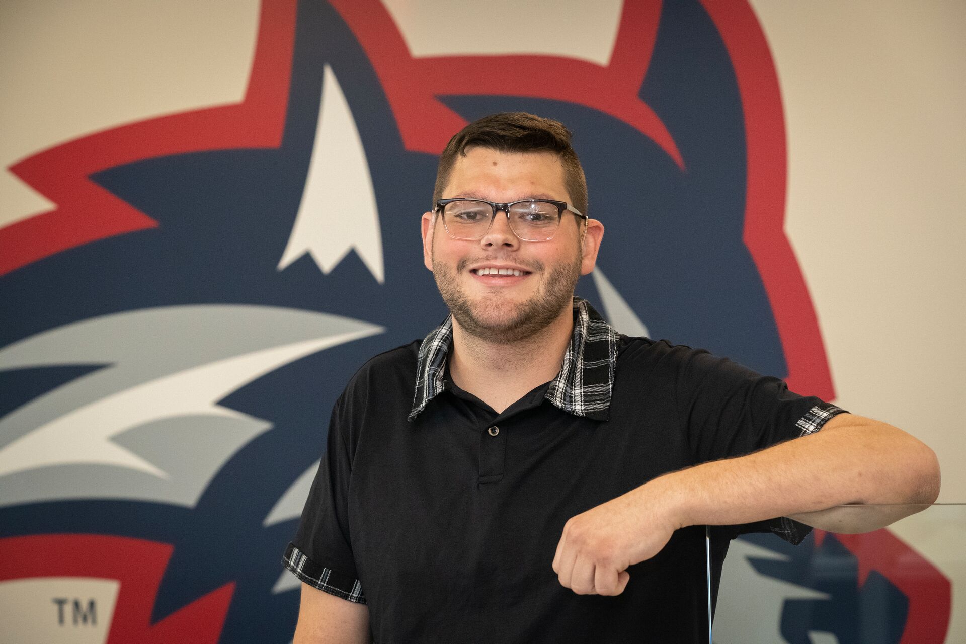 A man (Seth Vessa), with glasses and facial hair, wearing a black polo shirt with a plaid collar, smiling and leaning against a glass partition with a Stony Brook University wolfie logo on the wall in the background.