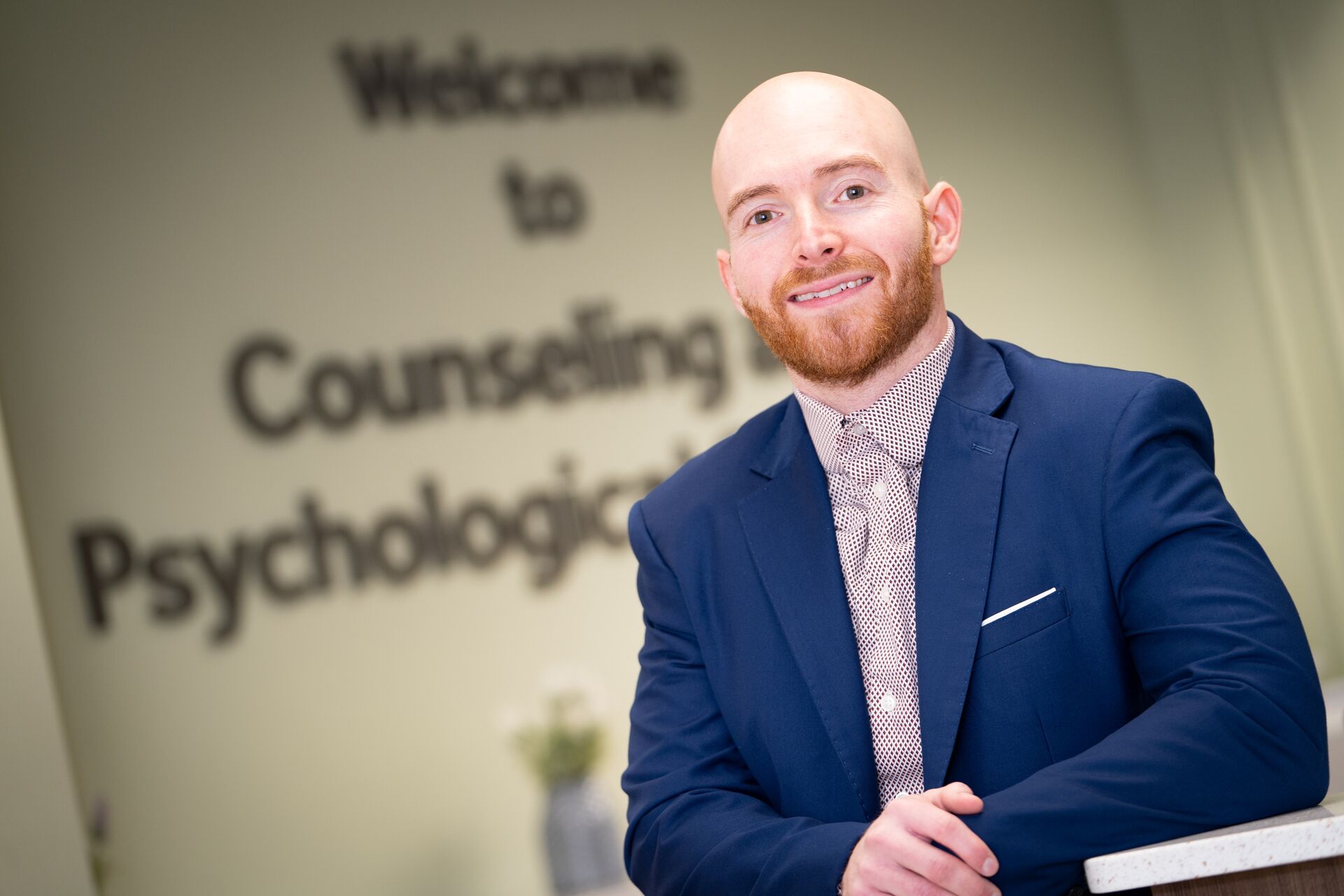 Caucasian man with shaved head and reddish facial hair, wearing a suit jacket and collared shirt smiling at the camera in front of a wall saying Welcome to Counseling and Psychological Services (Rob)