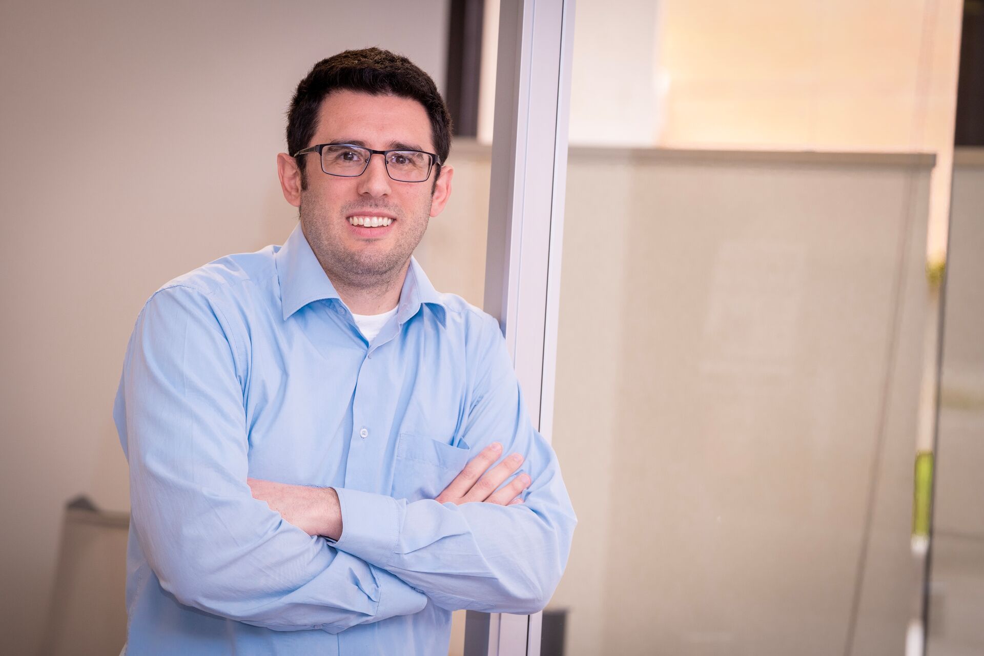 Young caucasian man with short black hair and glasses wearing a blue collared shirt and folding his arms smiling (Nick)