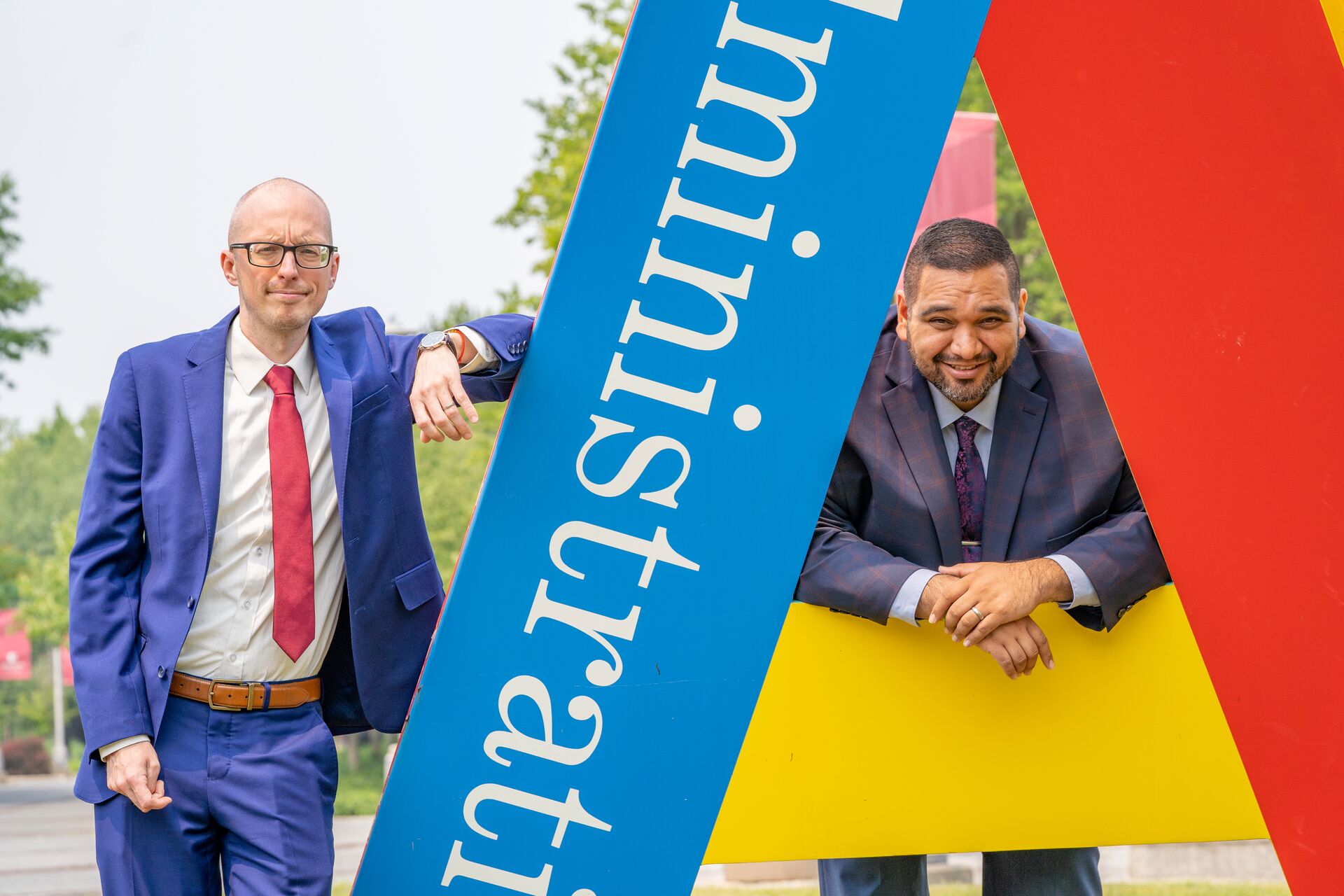 Mostly bald caucasian man with slight facial hair, wearing a blue suit, red tie and white shirt, leaning on the 'A' sculpture outside of the Admin Building, with another white male with a crew cut and facial hair and wearing a suit crouching inside the opening of the letter 'A' on his right