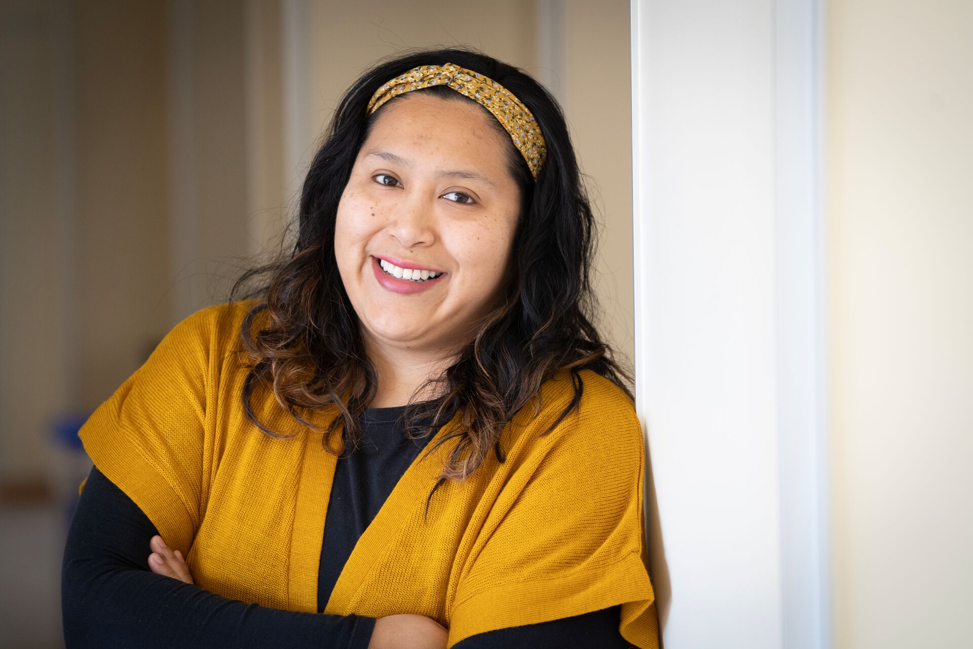 A woman (Janet Campoverde), with wavy dark hair and yellow headband leaning against a white wall wearing a yellow cardigan over a black top.