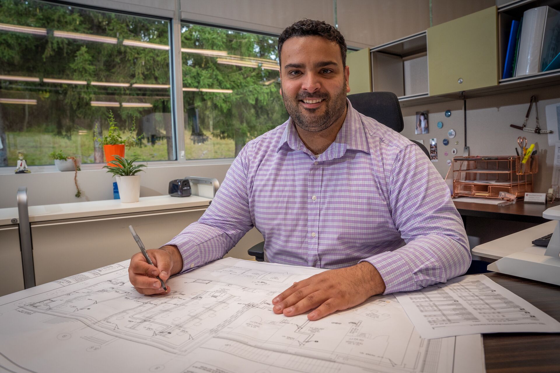 Caucasian man with facial hair and dress shirt seated, holding a pencil and working on architectural drawing on a desk