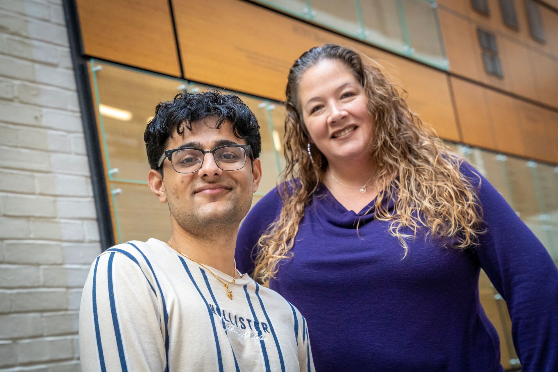 Young Indian male with dark short hair and glasses smiling with striped shirt (Krish) with long curly haired blonde white woman wearing a purple sweater (Maria) in front of a brick, glass and wood wall background 