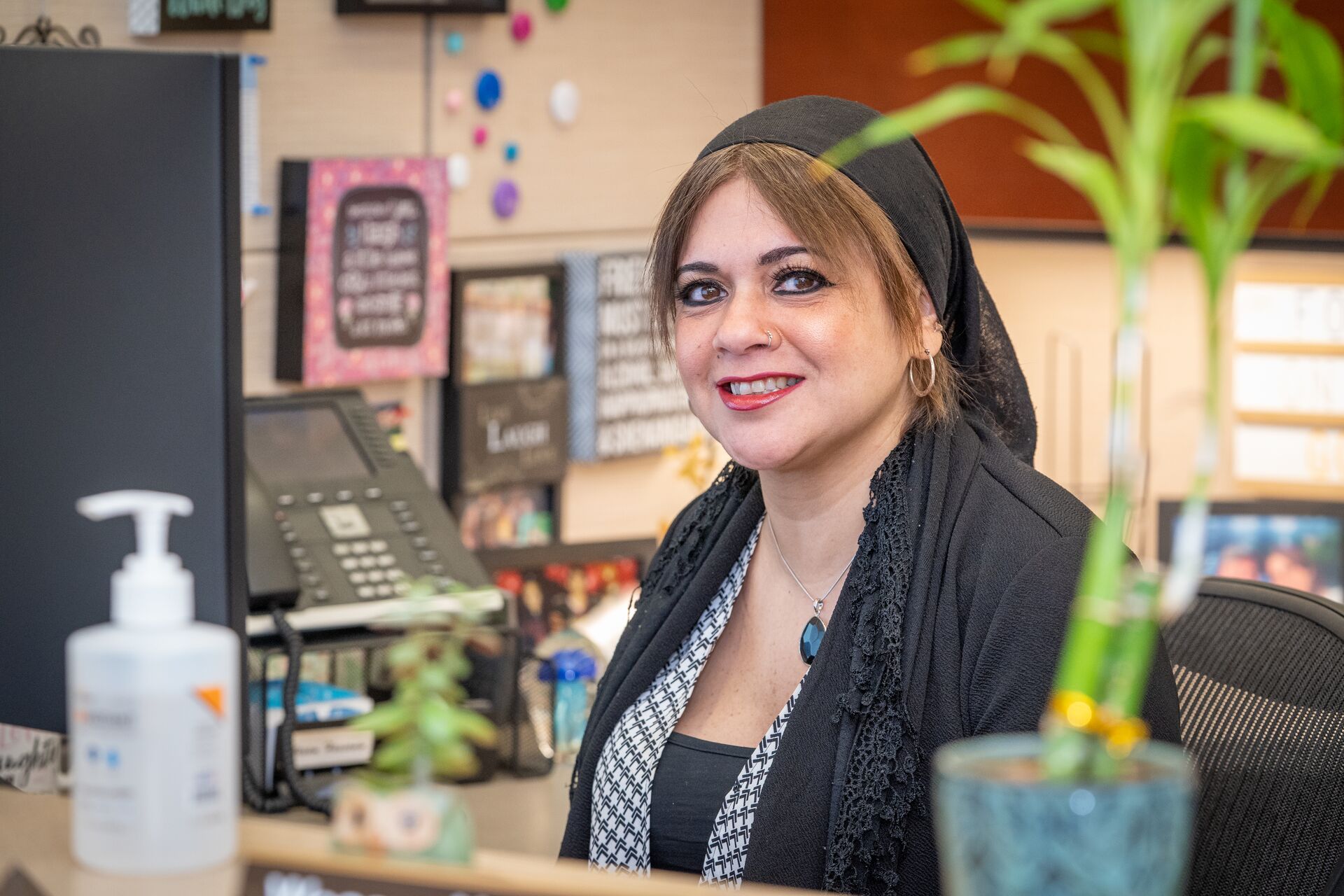 Woman with a hijab wearing and black jacket and black-and-white blouse smiling sitting by the computer at her desk in her office (Wesam)