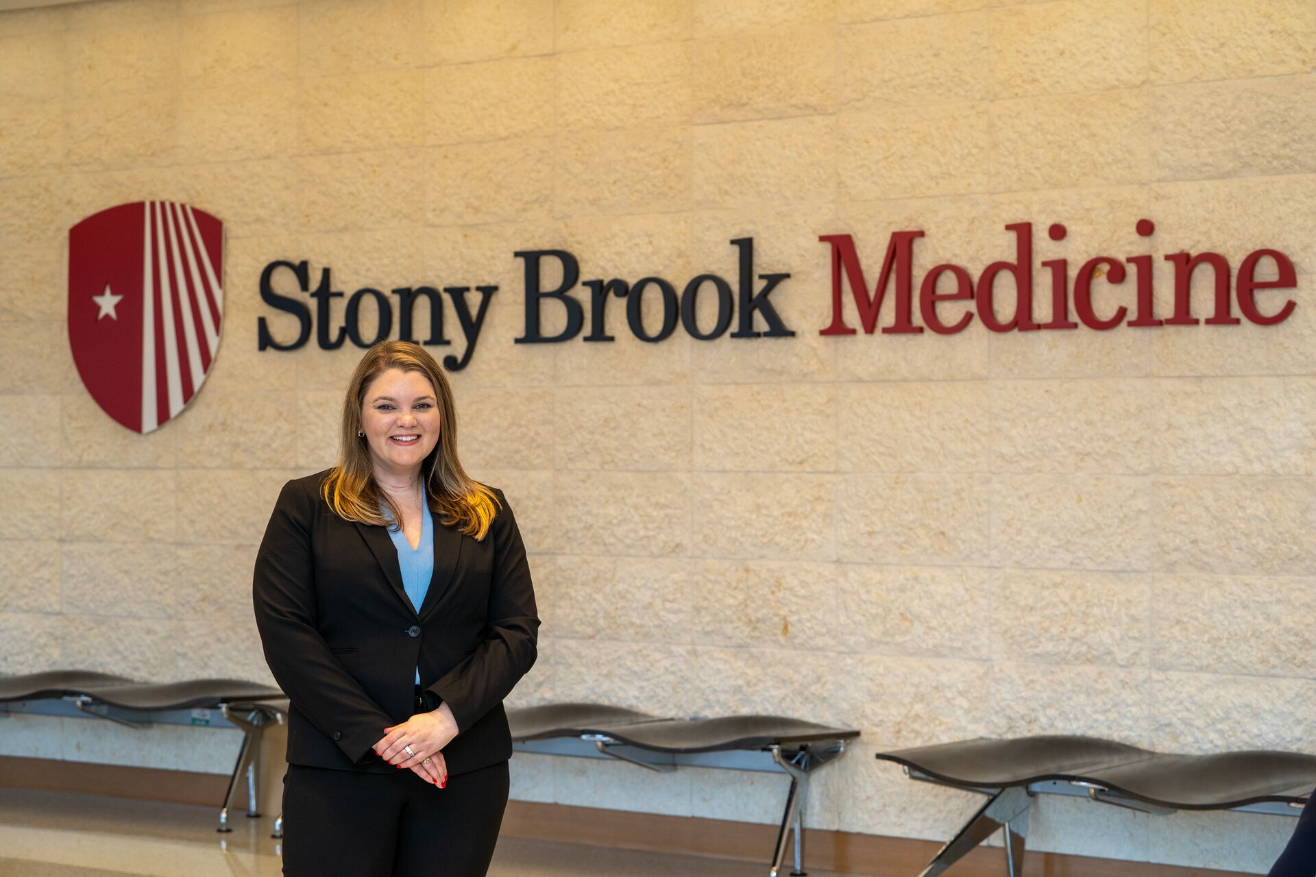 Young blonde caucasian woman in blue blazer and blouse standing smiling in front of a Stony Brook Medicine sign on the wall (Erika)
