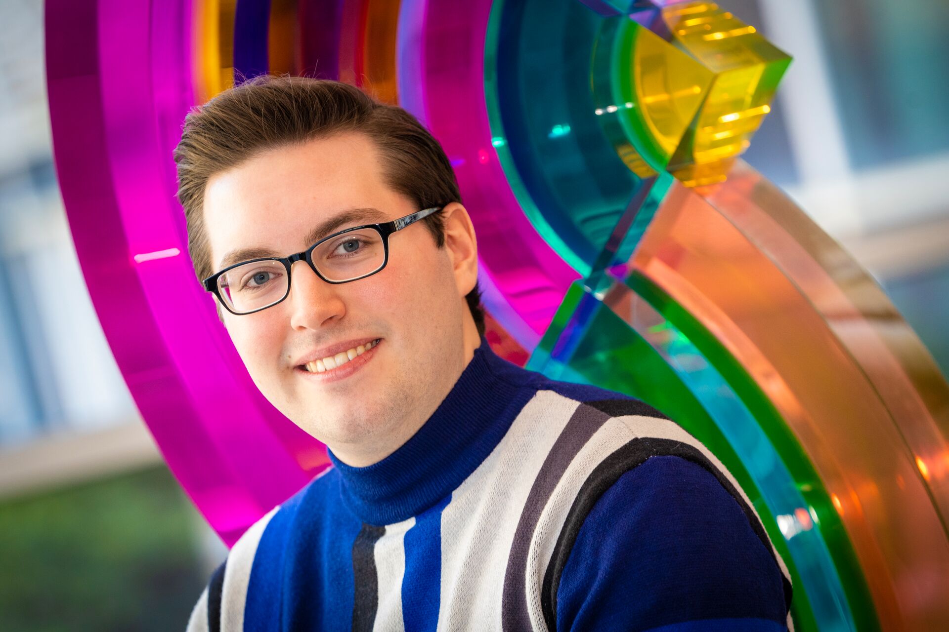 Young caucasian man with short brown hair and glasses smiling standing in the SAC lobby in front of the rainbow sculpture (Peter)