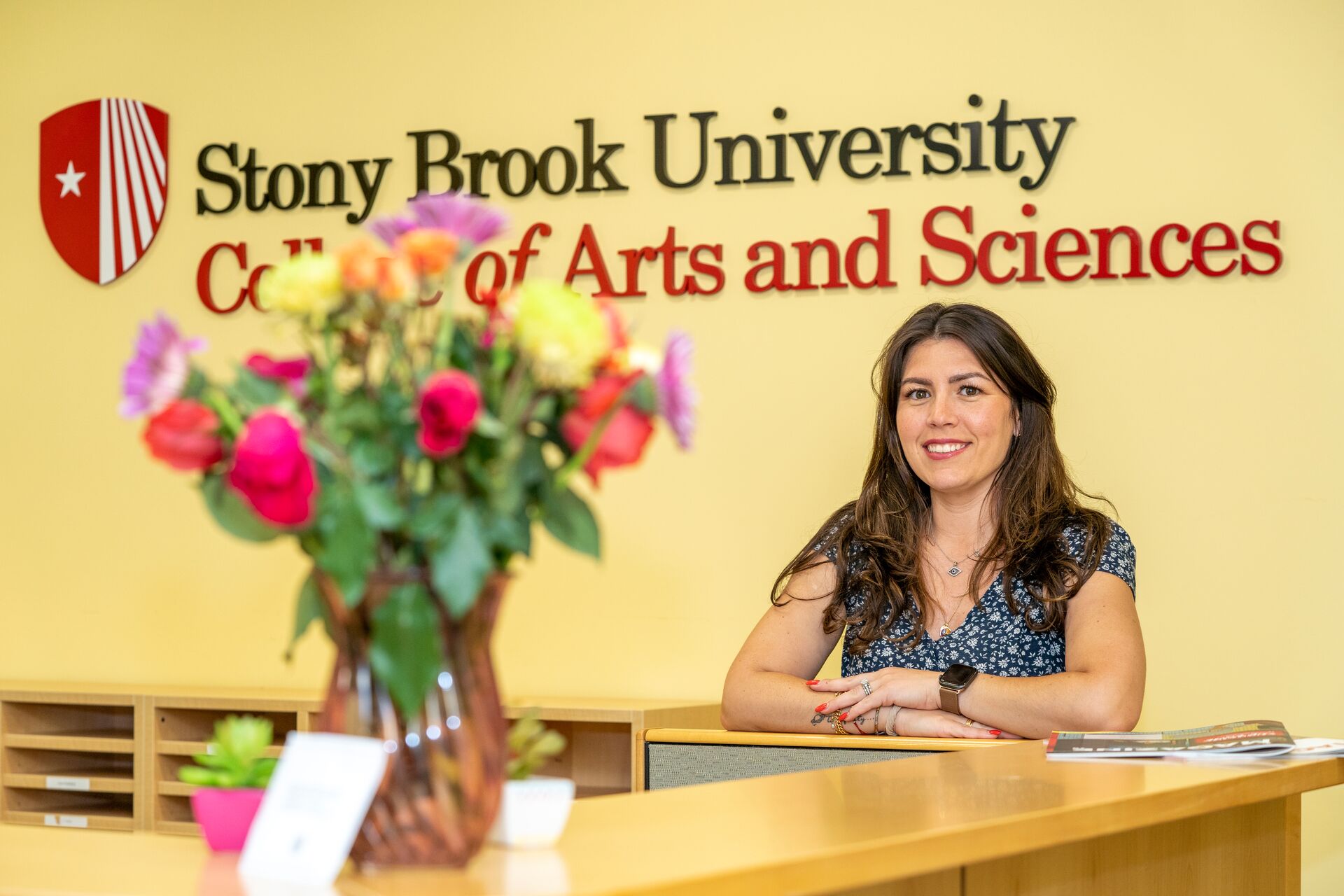 Young white woman with long dark brown hair (Danielle) wearing a flowery black blouse and smiling behind a desk with flowers and in front of a wall saying Stony Brook University College of Arts and Sciences