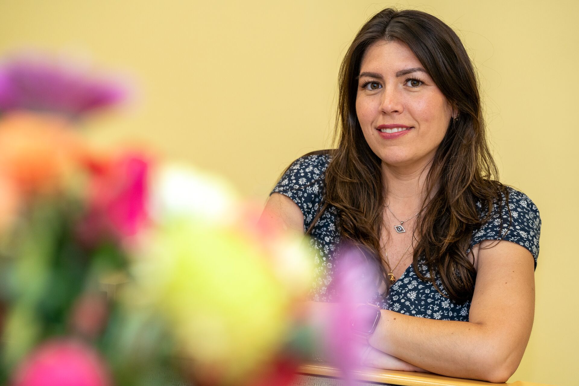 A woman (Danielle Belmonte), with long brown hair, wearing a navy blue floral top, smiling with her arms crossed. Colorful, blurred flowers are in the foreground against a yellow wall background.