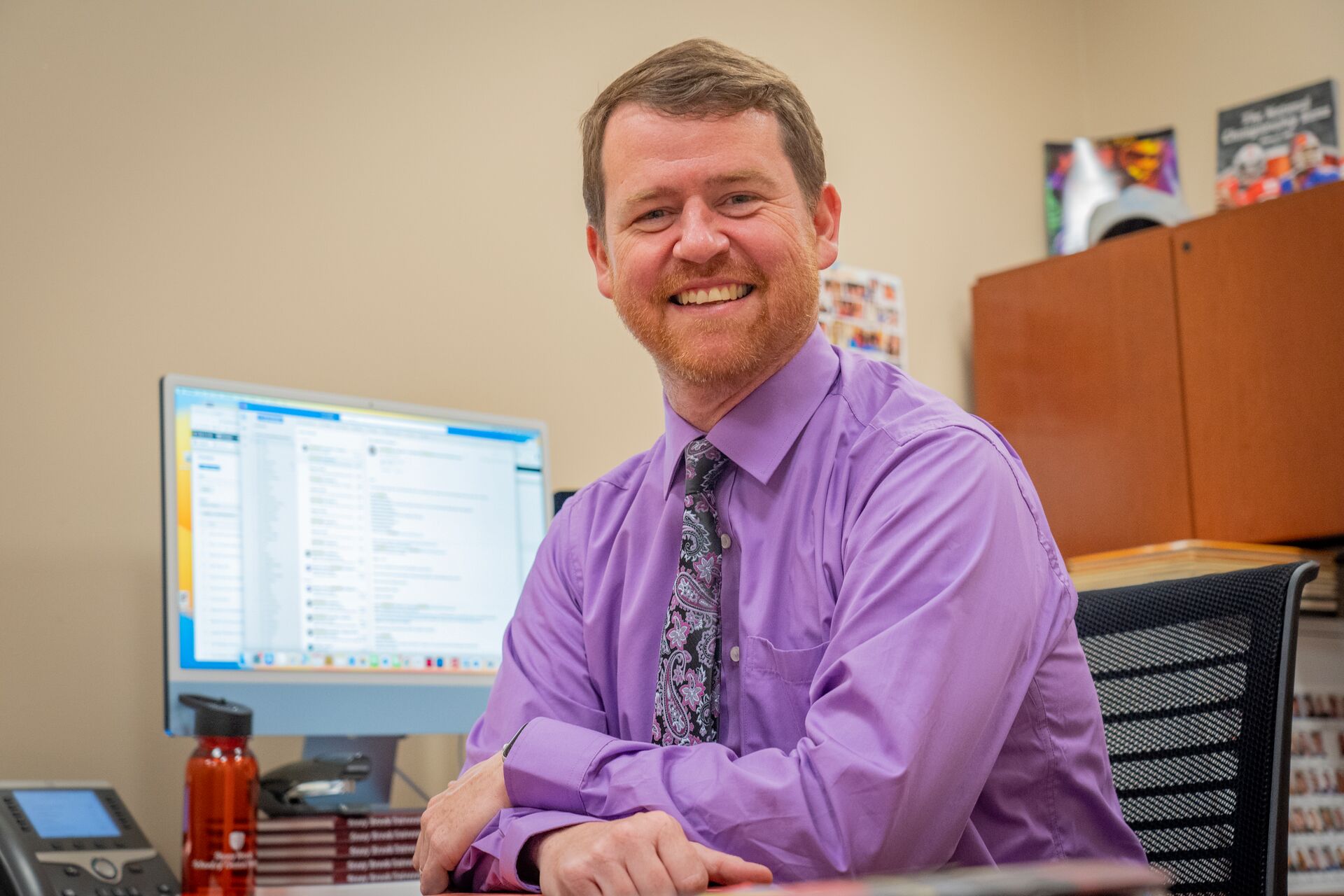 White male (Brett Mauser) with sandy-light brown hair and beard in a purple collared shirt and matching tie smiling at the camera in front of his computer