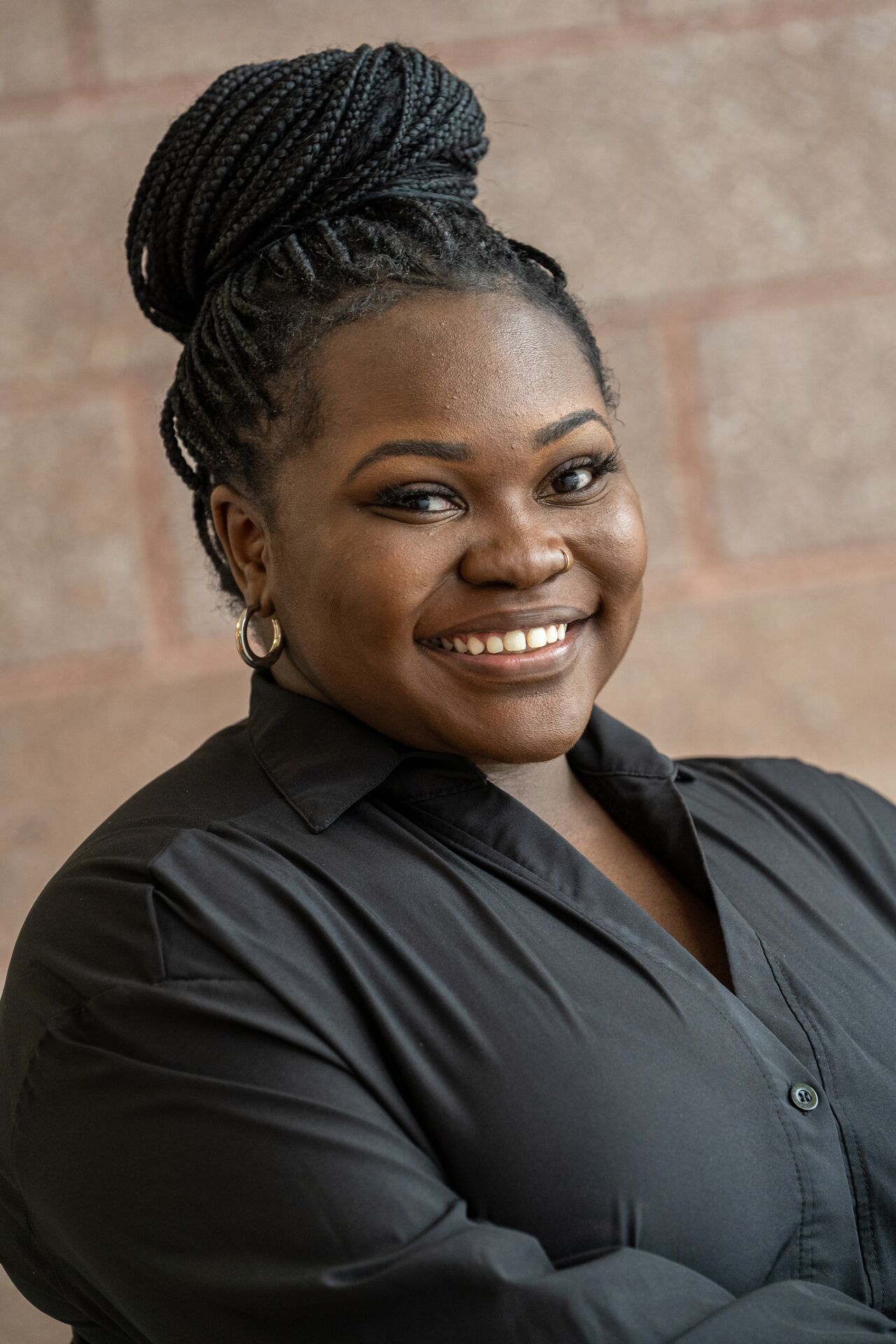 A woman (Natasha McCombs), with dark hair in a braided bun and wearing a black button-down shirt standing and smiling in front of a brick wall.