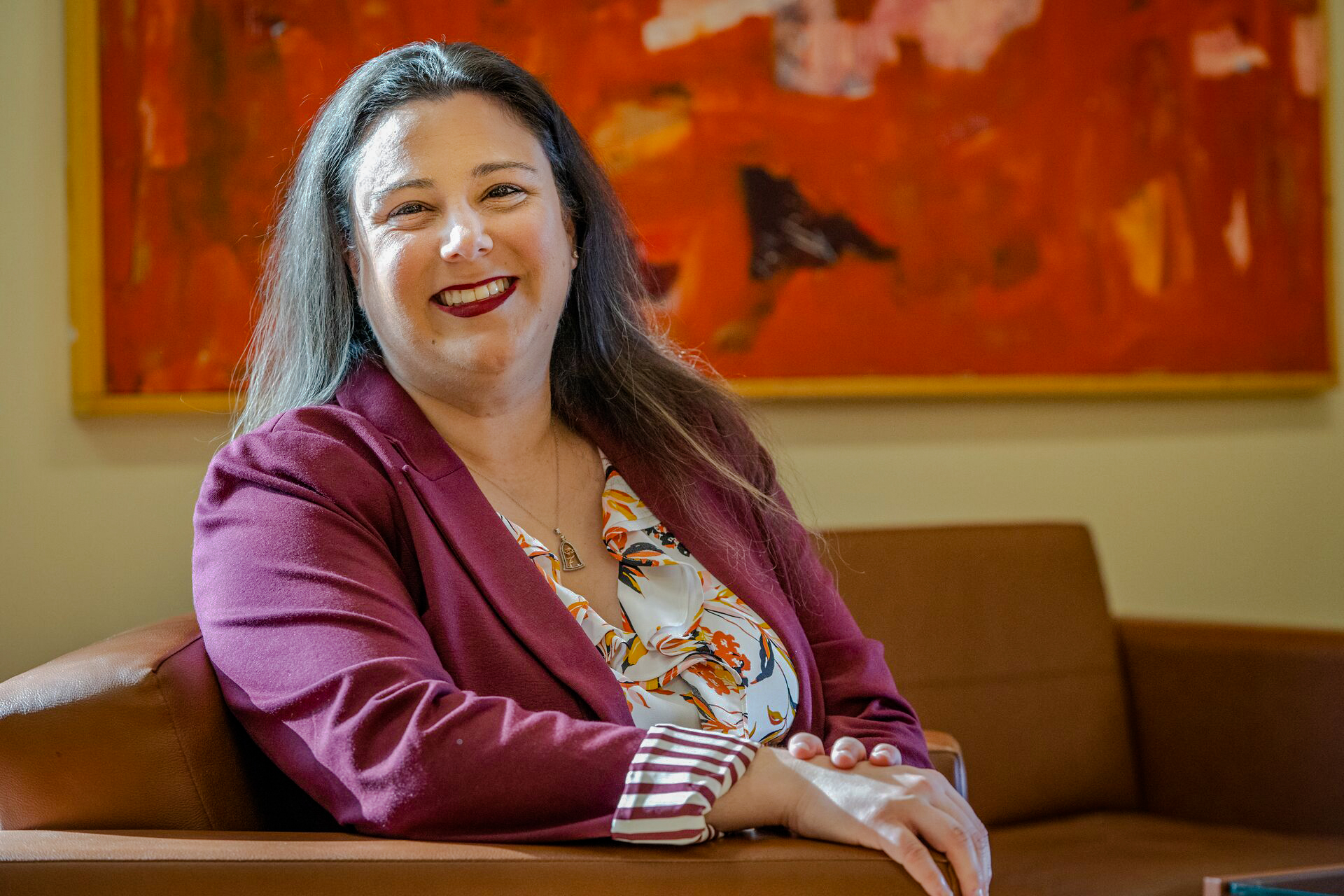 Causasian woman (Allison) with long brown hair, a pink blazer and white flowery blouse sitting and smiling on a couch in front of a colorful orange painting